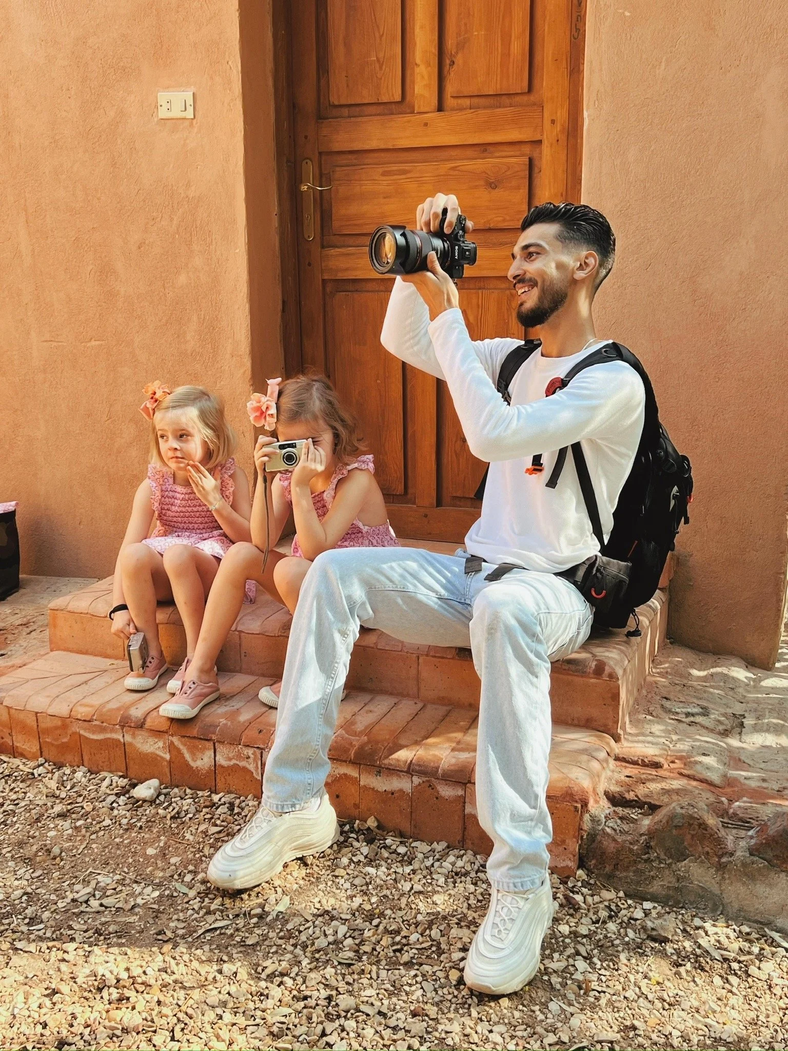 Omar, the photographer sitting on steps taking a photograph with a camera, accompanied by two young girls sitting next to him, one holding a camera and the other holding a smartphone, outside a building with a wooden door and beige walls.