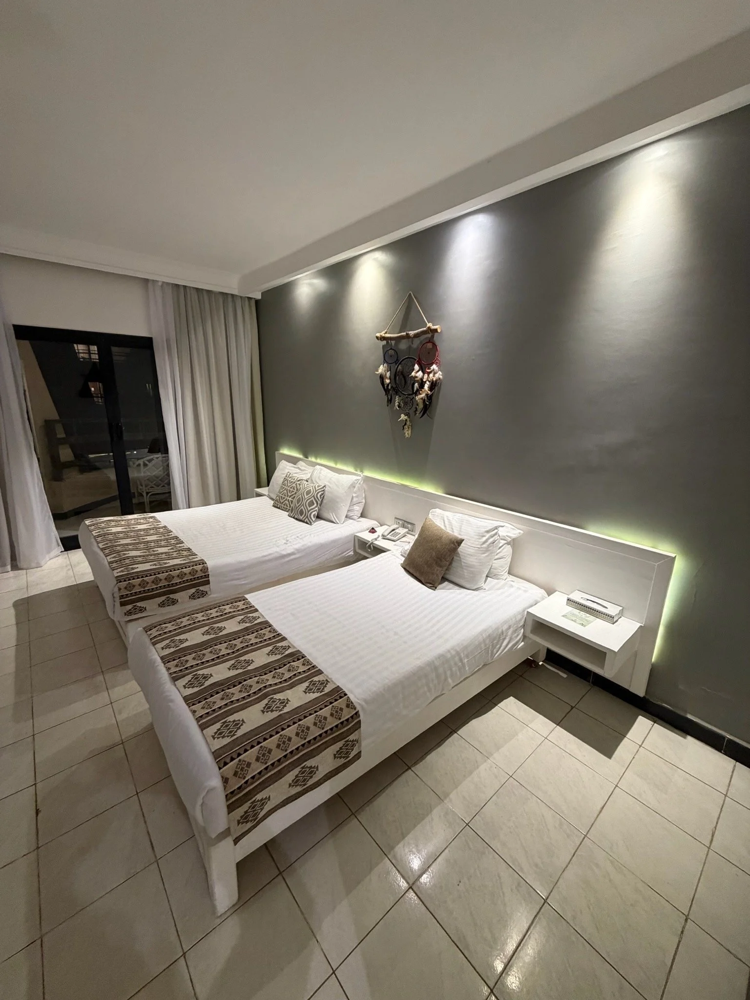 Two twin beds with white bedding and decorative pillows in a hotel room, featuring a gray accent wall with a dreamcatcher decoration and hanging lights above the beds.