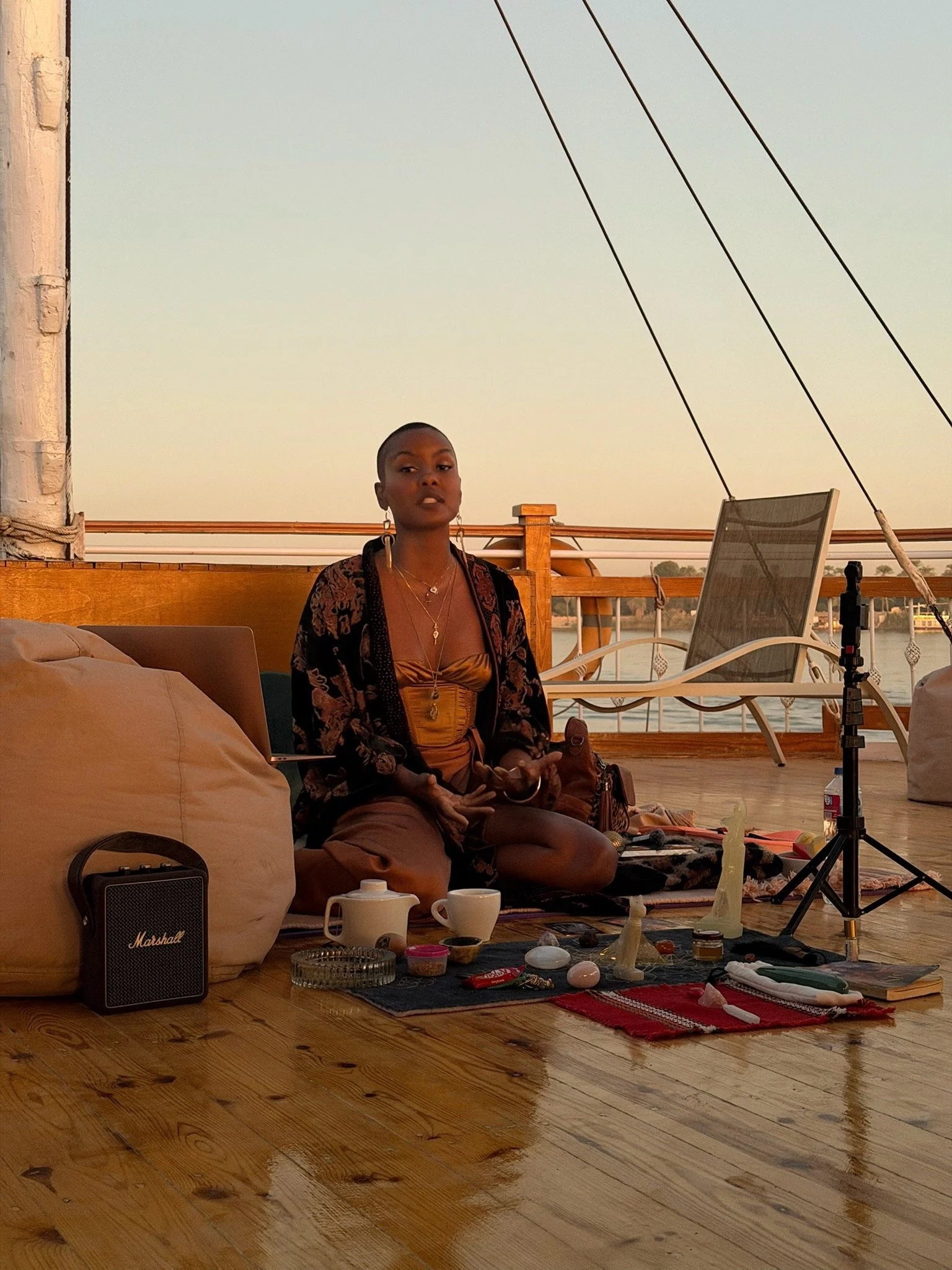 Andrea sitting cross-legged on the deck of the boat that takes you down the Nile, surrounded by various objects and equipment, with a large pillow, a speaker, and a laptop nearby, during sunset.