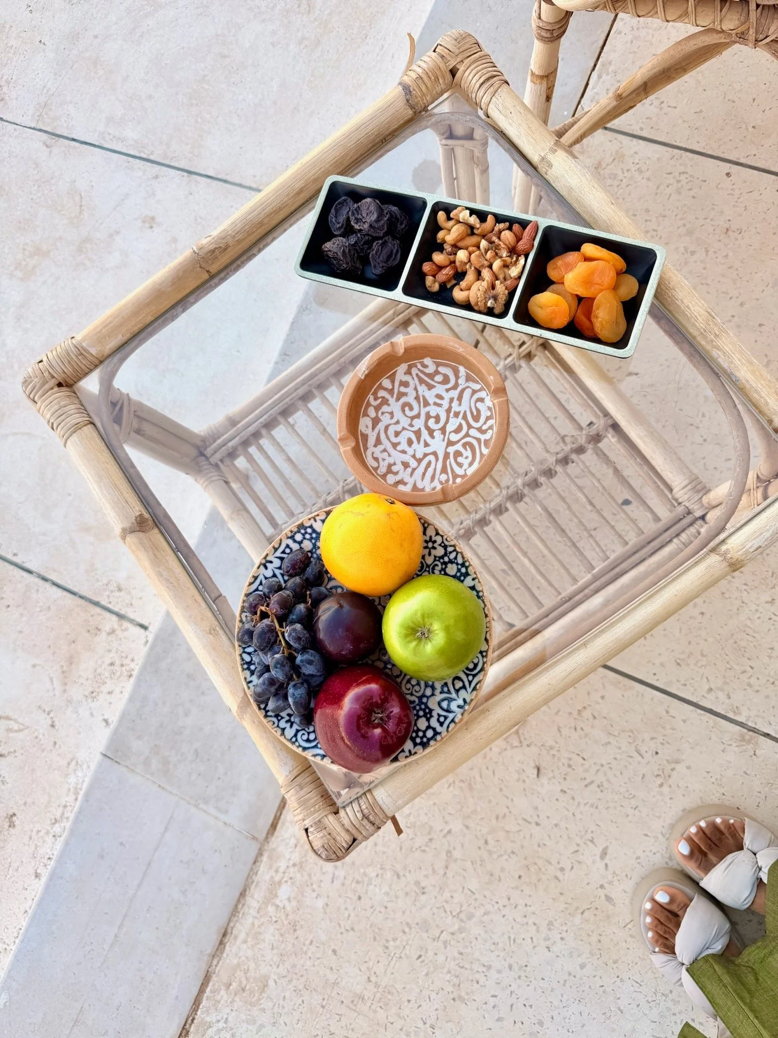 A glass-topped wicker table with a plate of grapes and apples, a small bowl of samosas, a rectangular dish with dried fruits and nuts, and a round bowl with a decorative pattern on a tile floor.