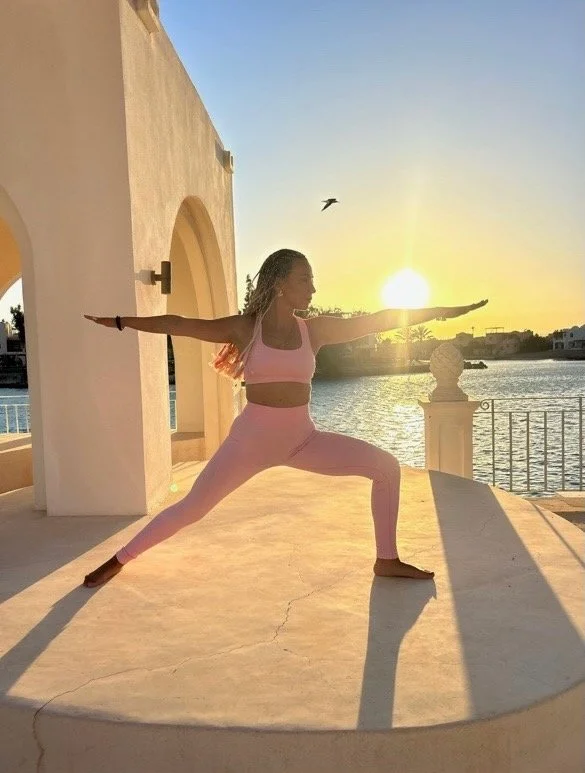Shereen practicing yoga in a warrior pose during sunrise by a waterfront, with a bird flying overhead and a sunny sky.