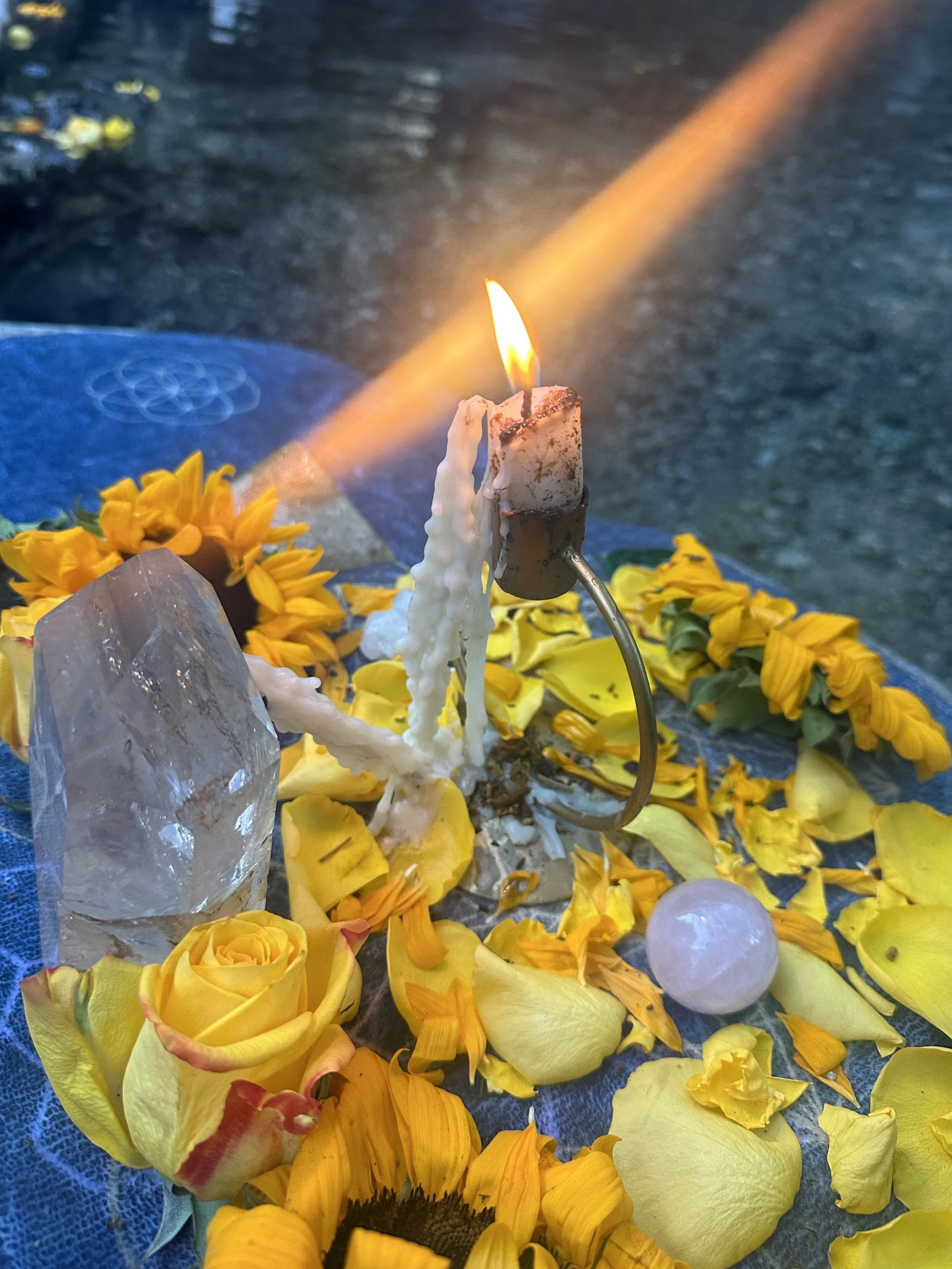 A memorial altar with yellow flowers, a lit candle, a large crystal, and a white spherical object, set against a mossy outdoor background.