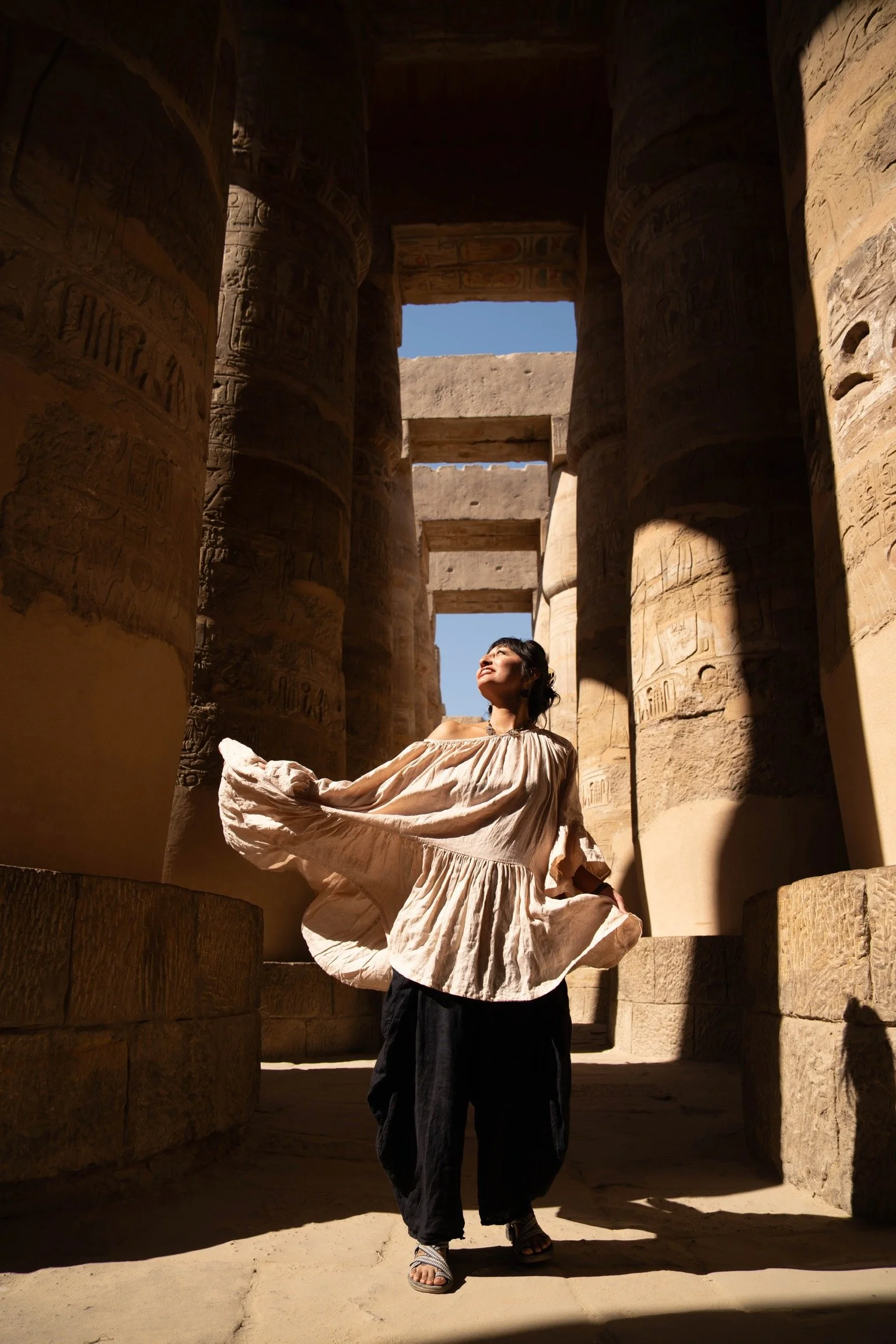 A woman in a flowing beige top and black pants stands in an ancient Egyptian temple with large stone columns decorated with hieroglyphics. She is looking up towards the sunlight streaming through the open top of the columns, with her right hand holding out her sleeve and her face illuminated by the sunlight.