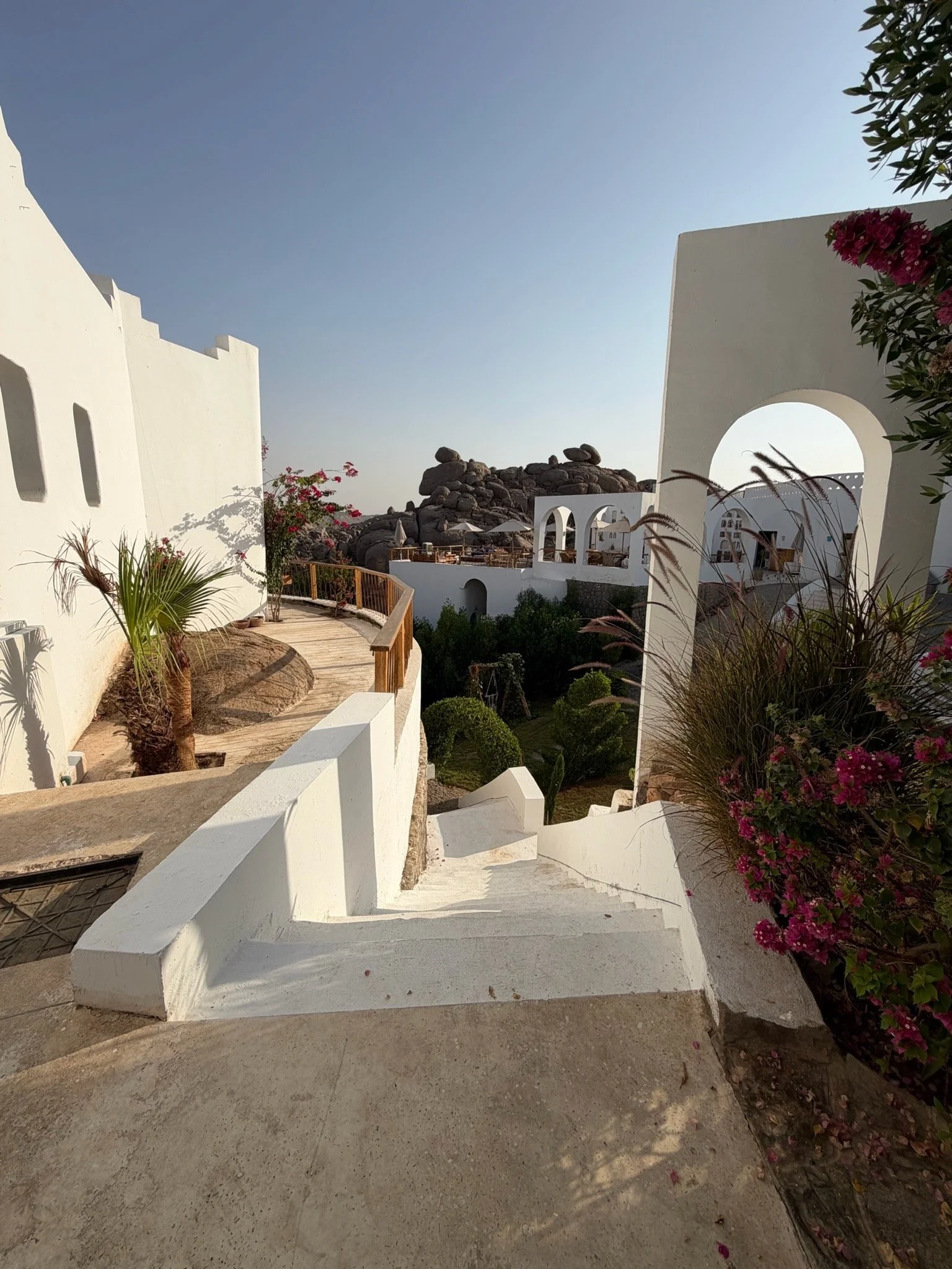 A scenic view of a Mediterranean-style white building complex with winding staircases, lush greenery, flowering plants, and rock formations in the background under a clear blue sky.