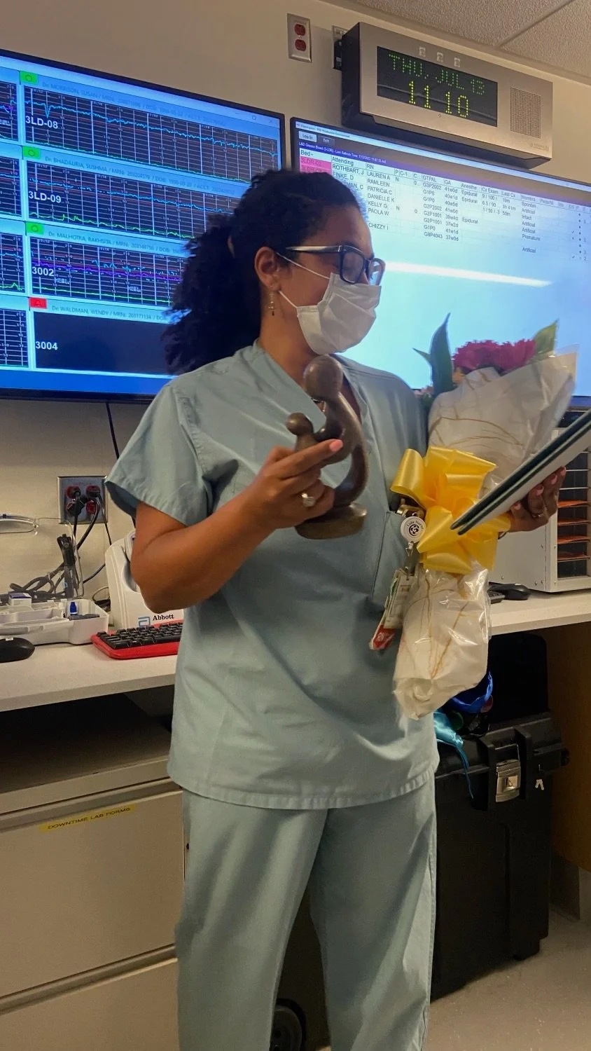 Nurse in scrubs and face mask holding a bouquet of flowers, a tablet, and a small statue, standing in a hospital room with monitoring screens in the background.