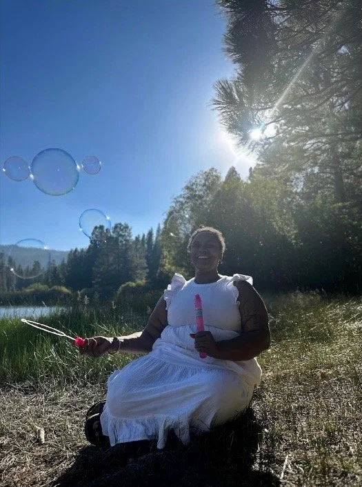 A woman wearing a white dress sitting outdoors near a body of water, blowing bubbles and enjoying the sunny day with trees and mountains in the background.