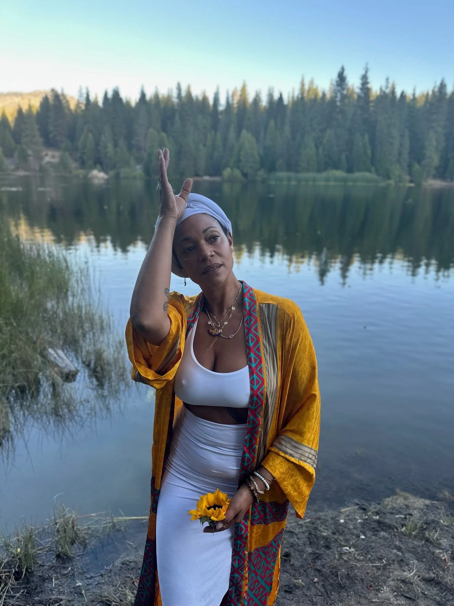 Paola standing by a lake with trees in the background. She is wearing a mustard yellow and colorful patterned outer garment, a white crop top, and white skirt. She has jewelry, a headwrap, and is holding a sunflower.