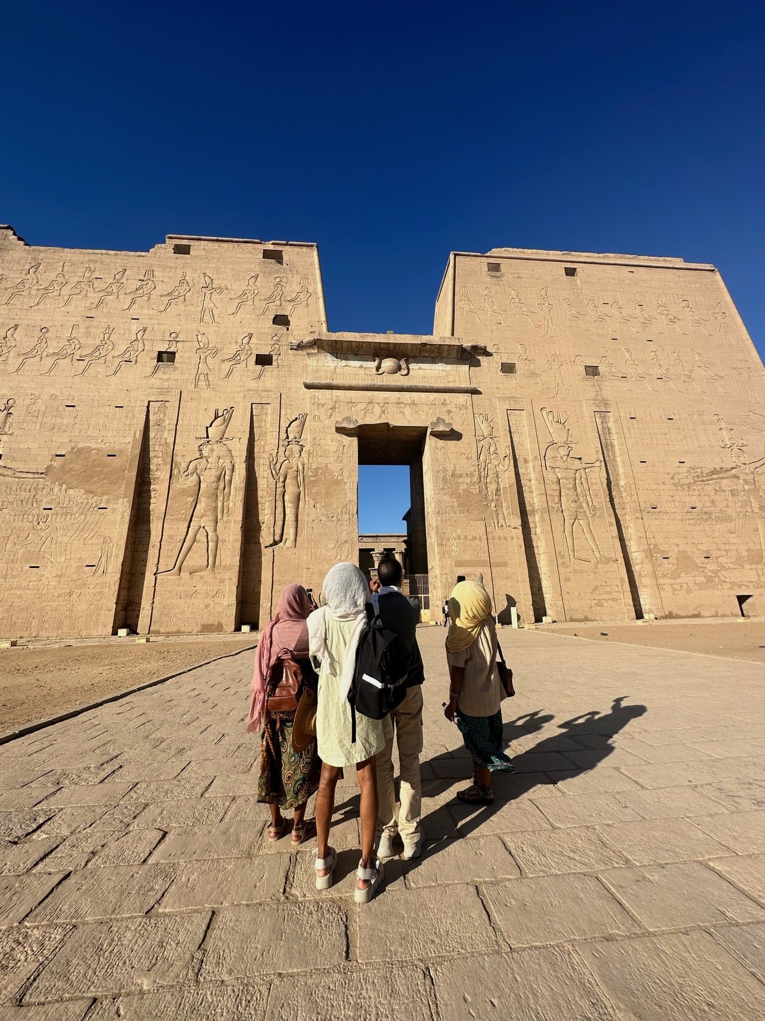 Tourists walking towards the entrance of an ancient Egyptian temple, with tall stone walls engraved with hieroglyphs and carvings, under a clear blue sky.