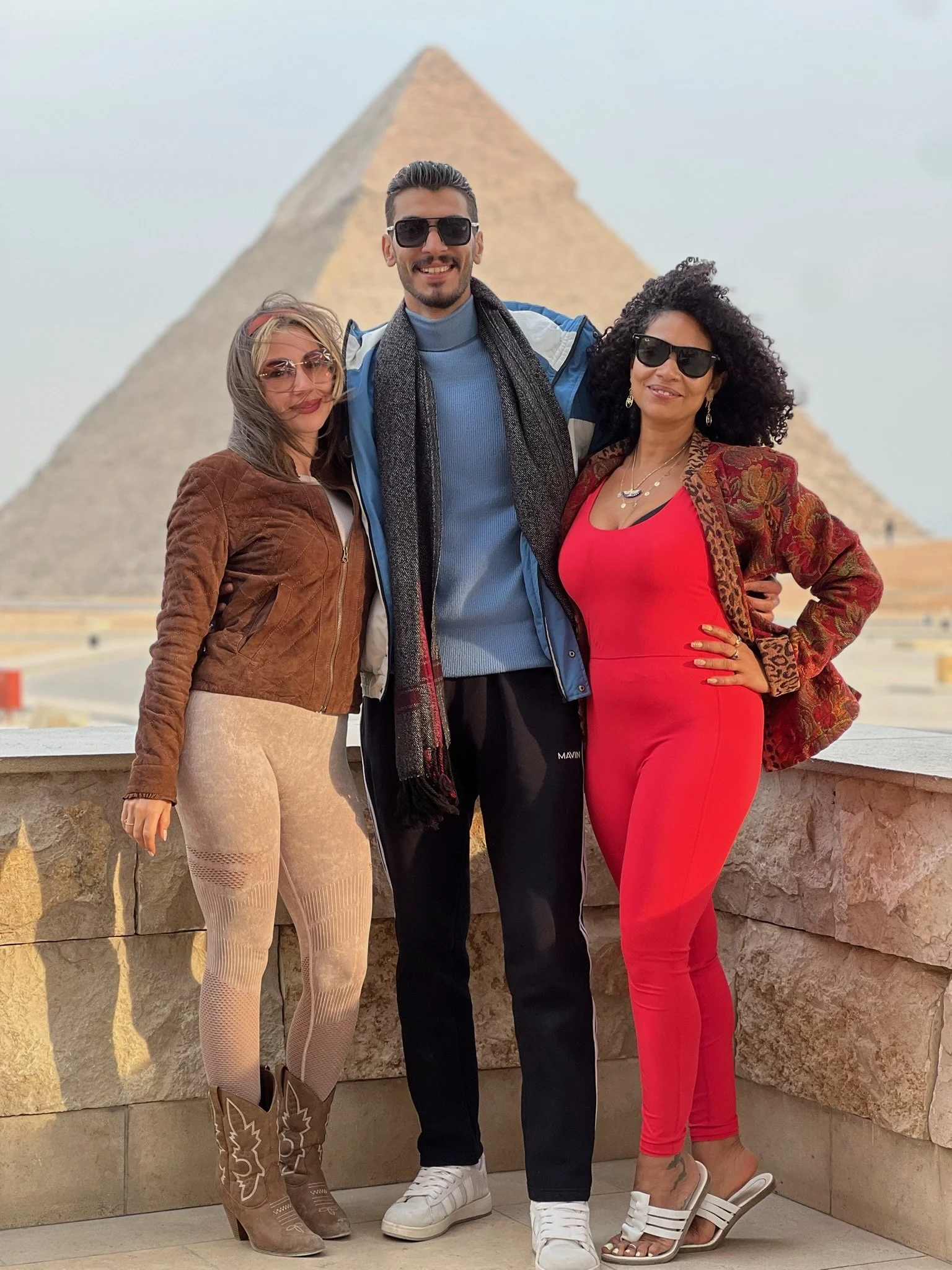 Shereen, Paola and Omar standing in front of the Great Pyramid of Giza, smiling for the camera, wearing sunglasses and fashionable clothing.