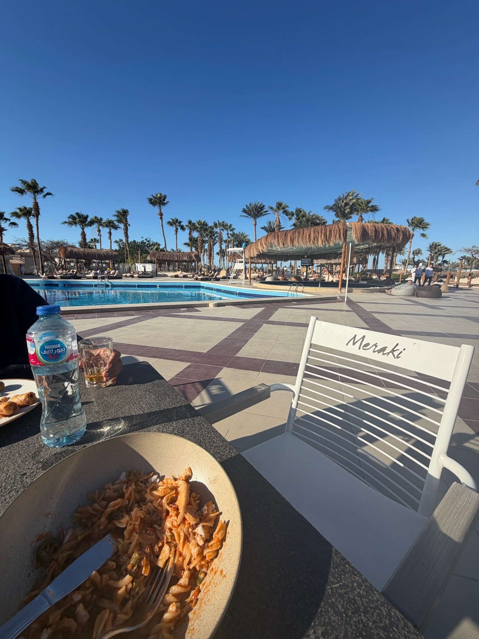 A poolside dining area at a resort with a large swimming pool, palm trees, thatched umbrellas, and lounge chairs under a clear blue sky. In the foreground, there is a table with a plate of pasta, a bottle of water, and a glass. A white chair with the