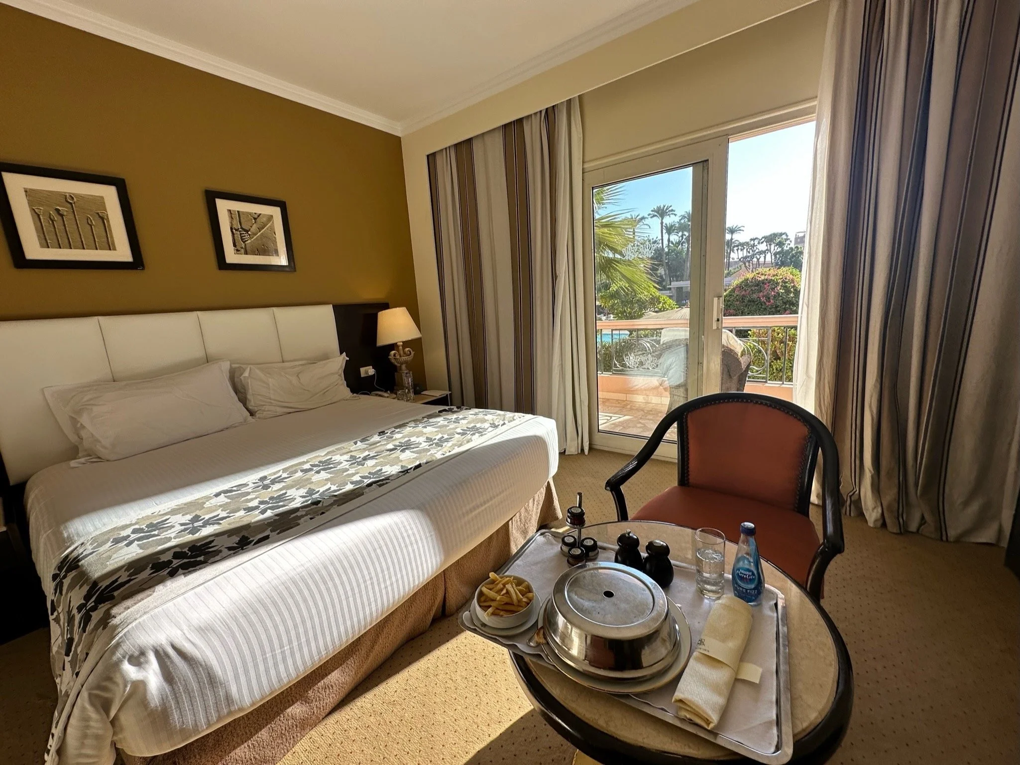 Hotel room with a bed, bedside lamp, pictures on the wall, sliding glass door leading to a balcony with outdoor furniture, and a table with a covered dish, French fries, water, and condiments.