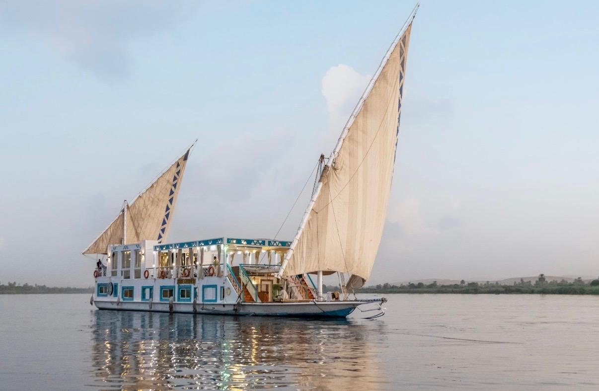 A large boat with beige sails on calm water, with a blue and white structure for passengers, and a shoreline with trees in the distance.