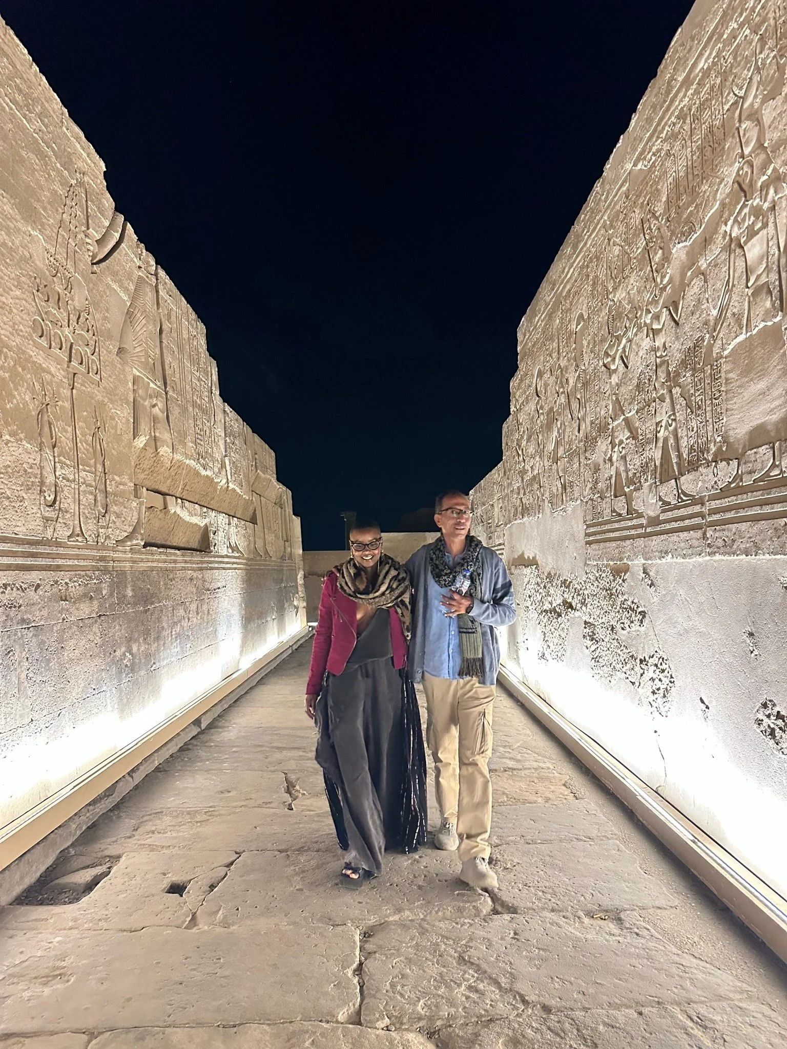 Andrea and Hassan, the Kemetikogist,  walking through an ancient Egyptian temple corridor with hieroglyphics carved into the walls, illuminated at night.