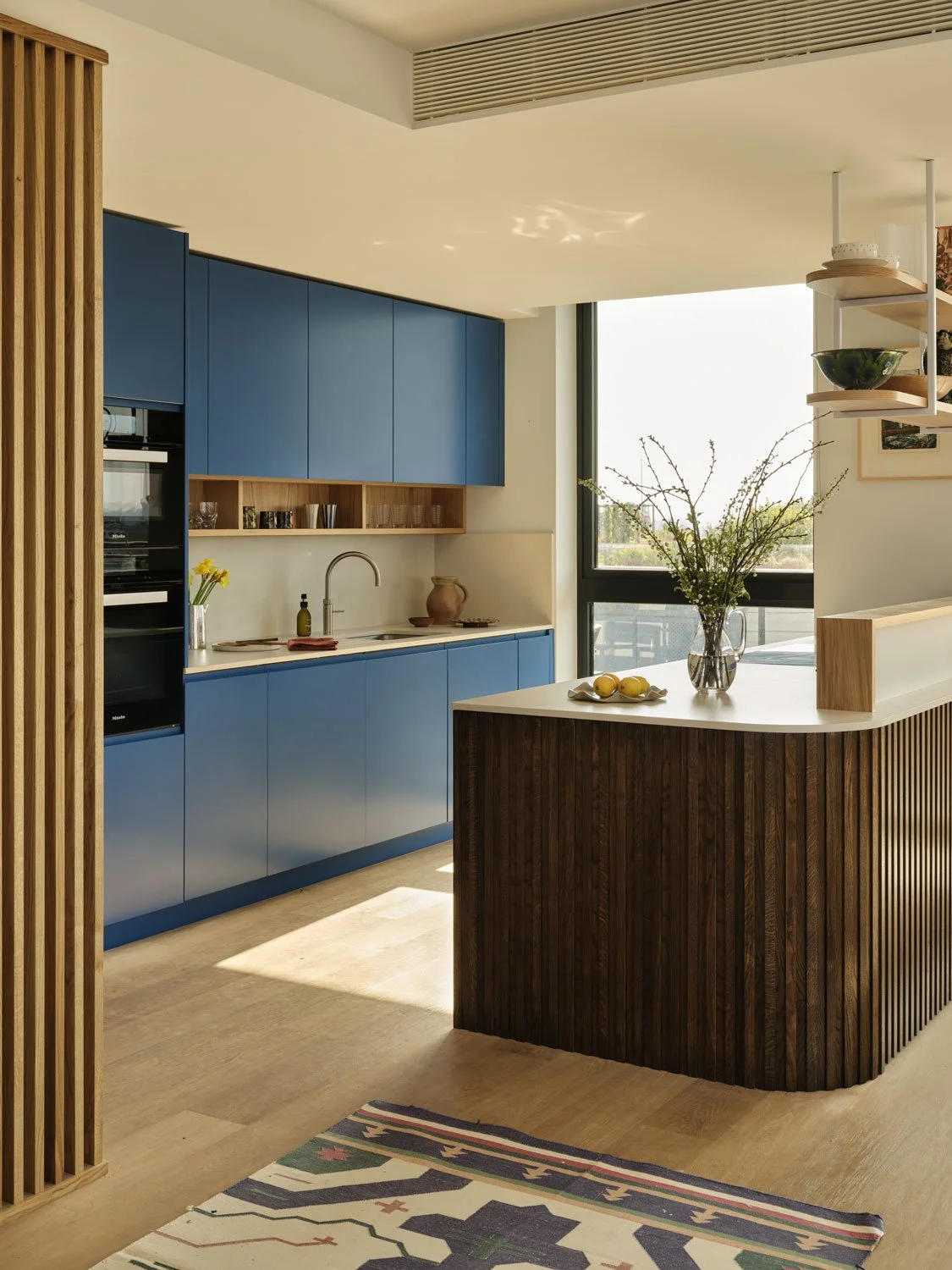 A bold blue kitchen with curved slatted oak island. A warm white quartz worktop creates a clean and calm feel. Suspended shelving above the island allow display of beautiful ceramics
