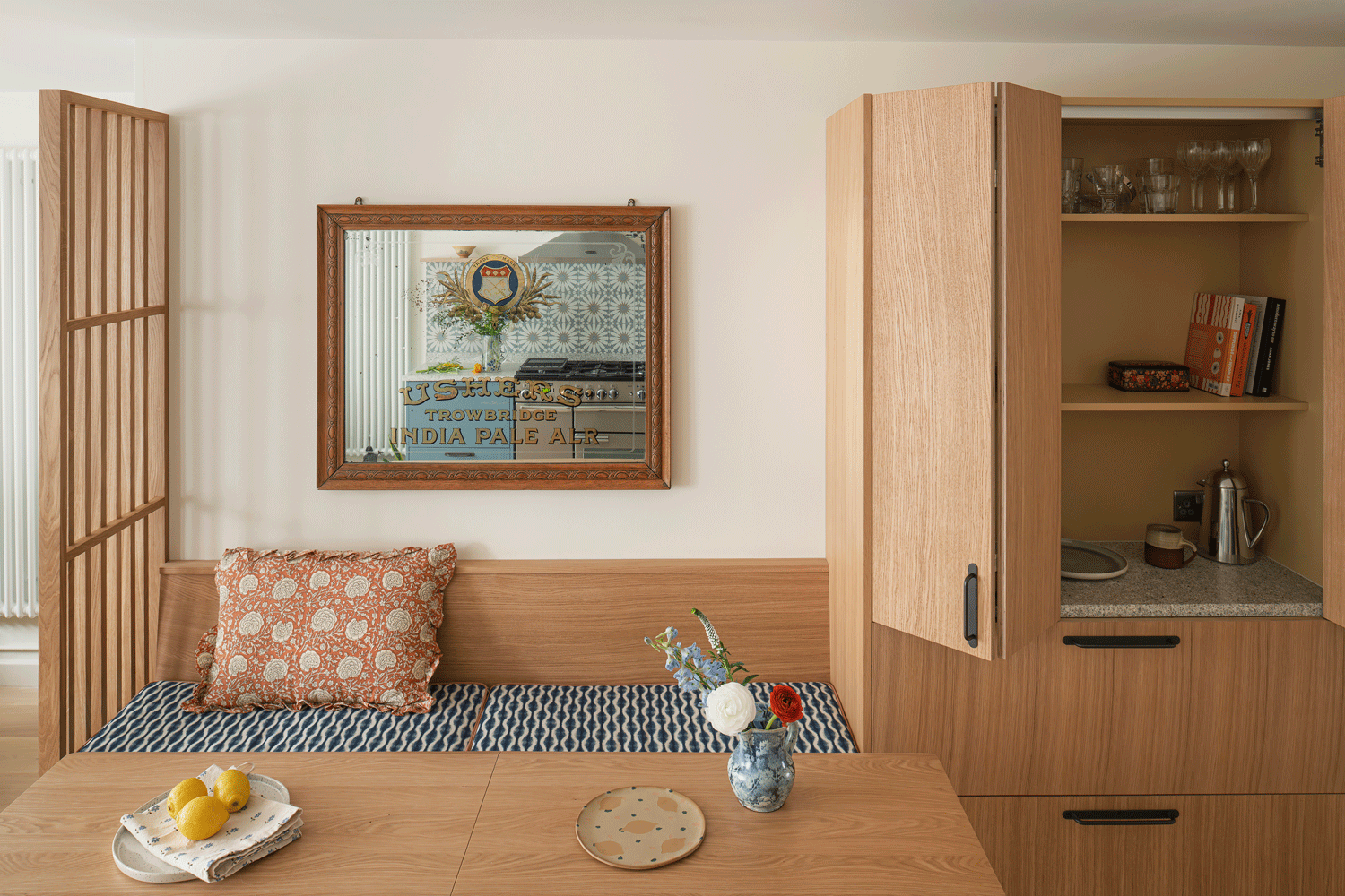 An oak dining bench and table is positioned between an oak slatted screen and bespoke made pantry unit. Bold fabric cushions add softness and pretty pattern