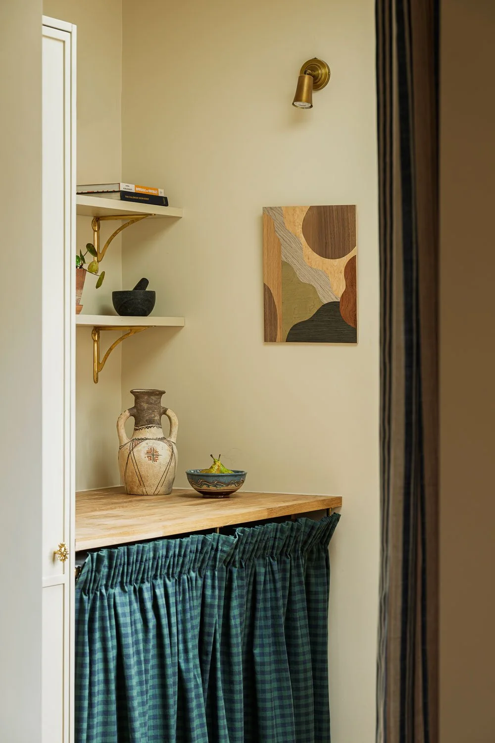 A contemporary kitchen in Knowle Bristol showing the view through to the utility space with open shelving on brass brackets and a cheque cafe curtain to hide the utilities. A unique bold artwork sits under an antique brass picture light