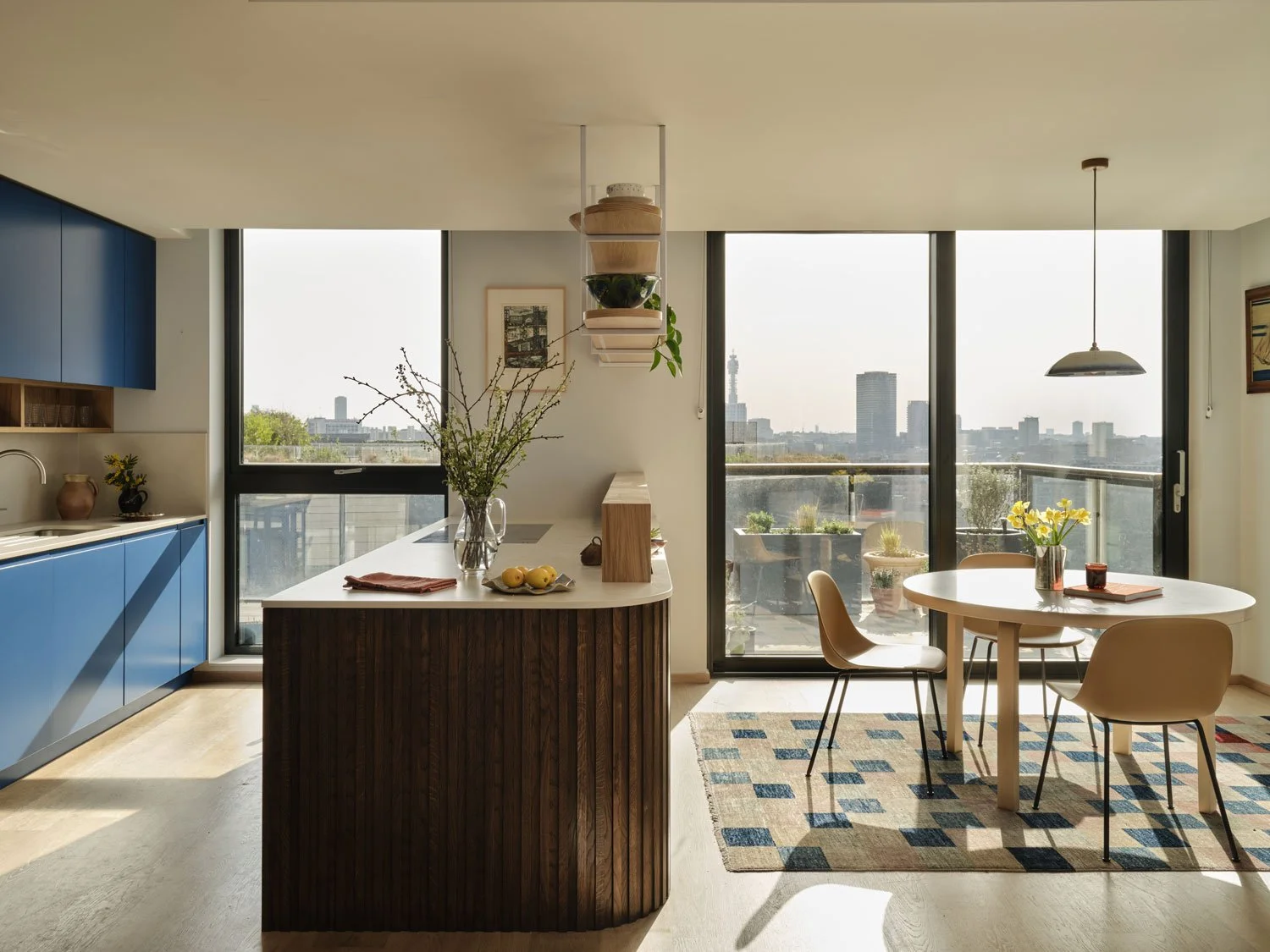 A view of a contemporary bold blue kitchen with curved oak slatted island. Large windows show views of King's Cross in London. A round dining table is grounded by a patterned rug and Studio Haran pendant light