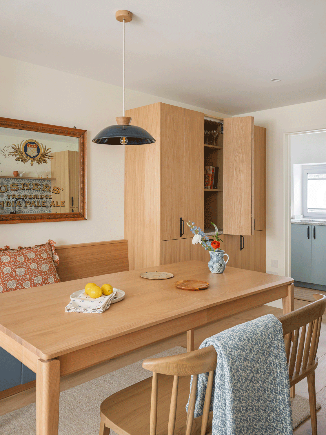 An oak dining table and integrated bench and pantry unit with a dark blue ceramic pendant. Pretty floral patterned soft furnishings add subtle pretty pattern