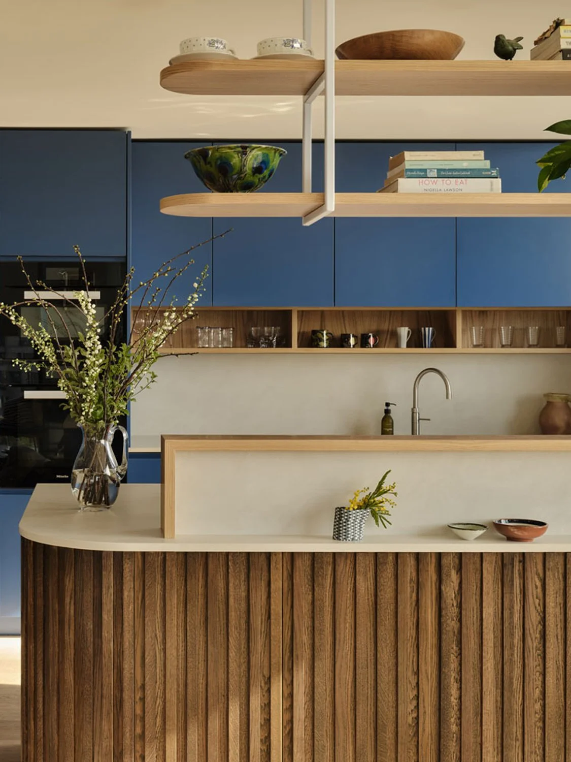 A detail of a stained oak slatted kitchen island and bold blue cabinetry. Suspended oak shelving allow for display of kitchen items