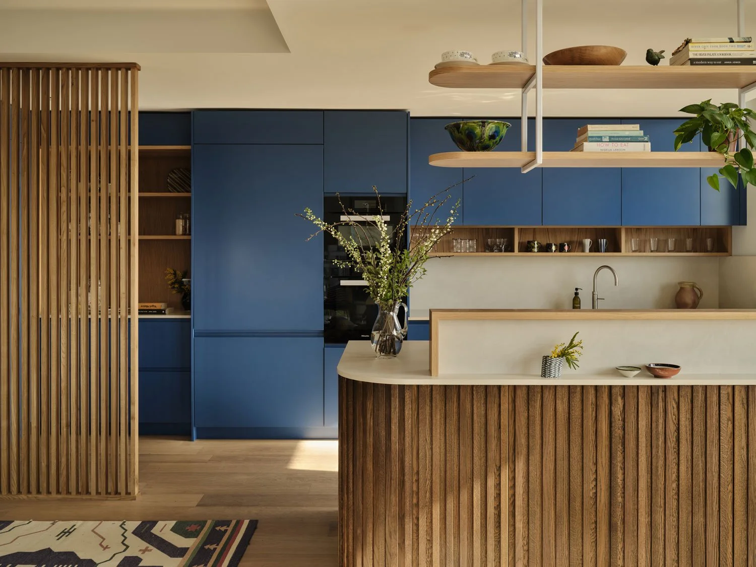 A bold blue modern kitchen with warm oak slatted island. Suspended oak shelves allow for the display of treasured items. A clean and contemporary kitchen with bold colour and warmth brought in with natural materials