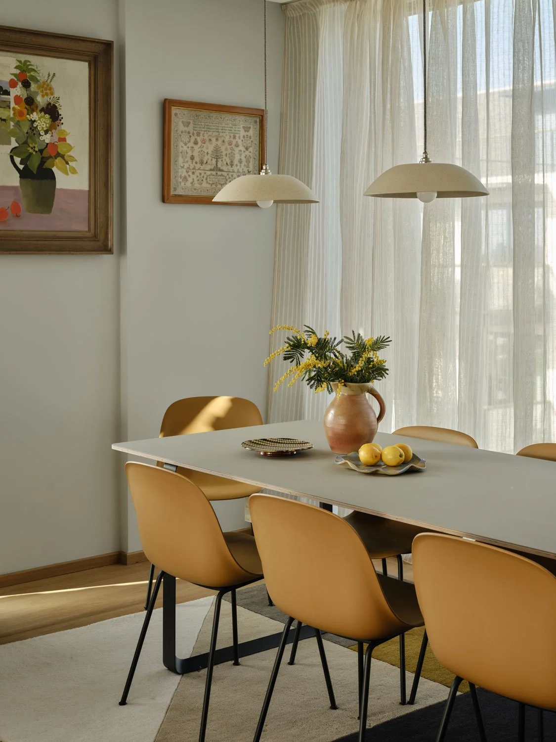A large contemporary dining table with soft yellow modern dining chairs. Studio Haran pendants hang low over the table. Colourful artwork and floaty linen drapes create a welcoming feel