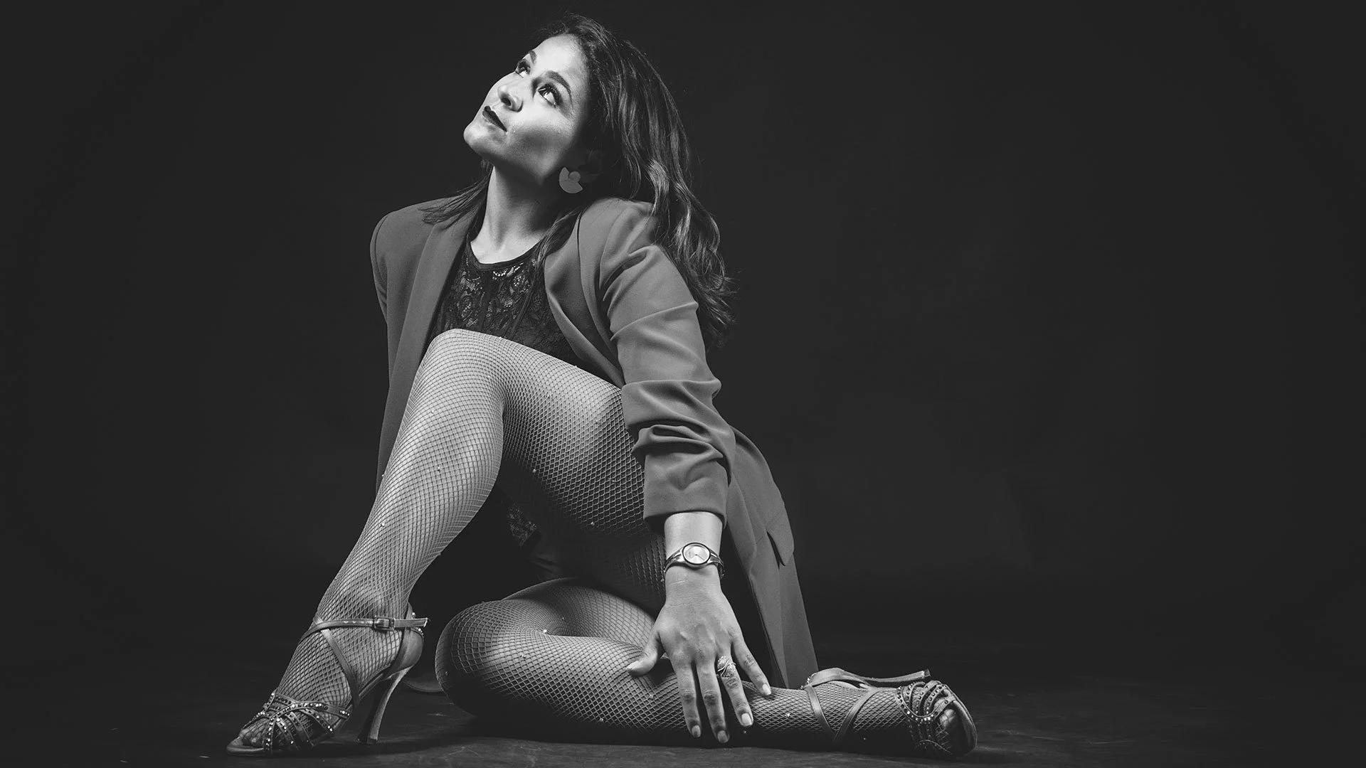 Mujer en pose artística, usando vestimenta elegante y medias de rejilla, en un fondo negro.