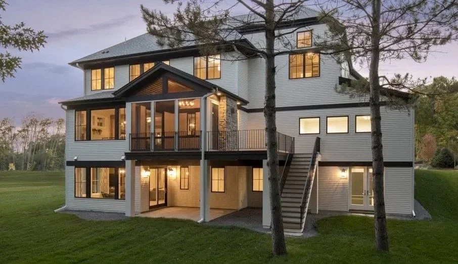 Large modern house with multiple stories, balcony, outdoor stairs, and illuminated windows, set on a grassy landscape at dusk.