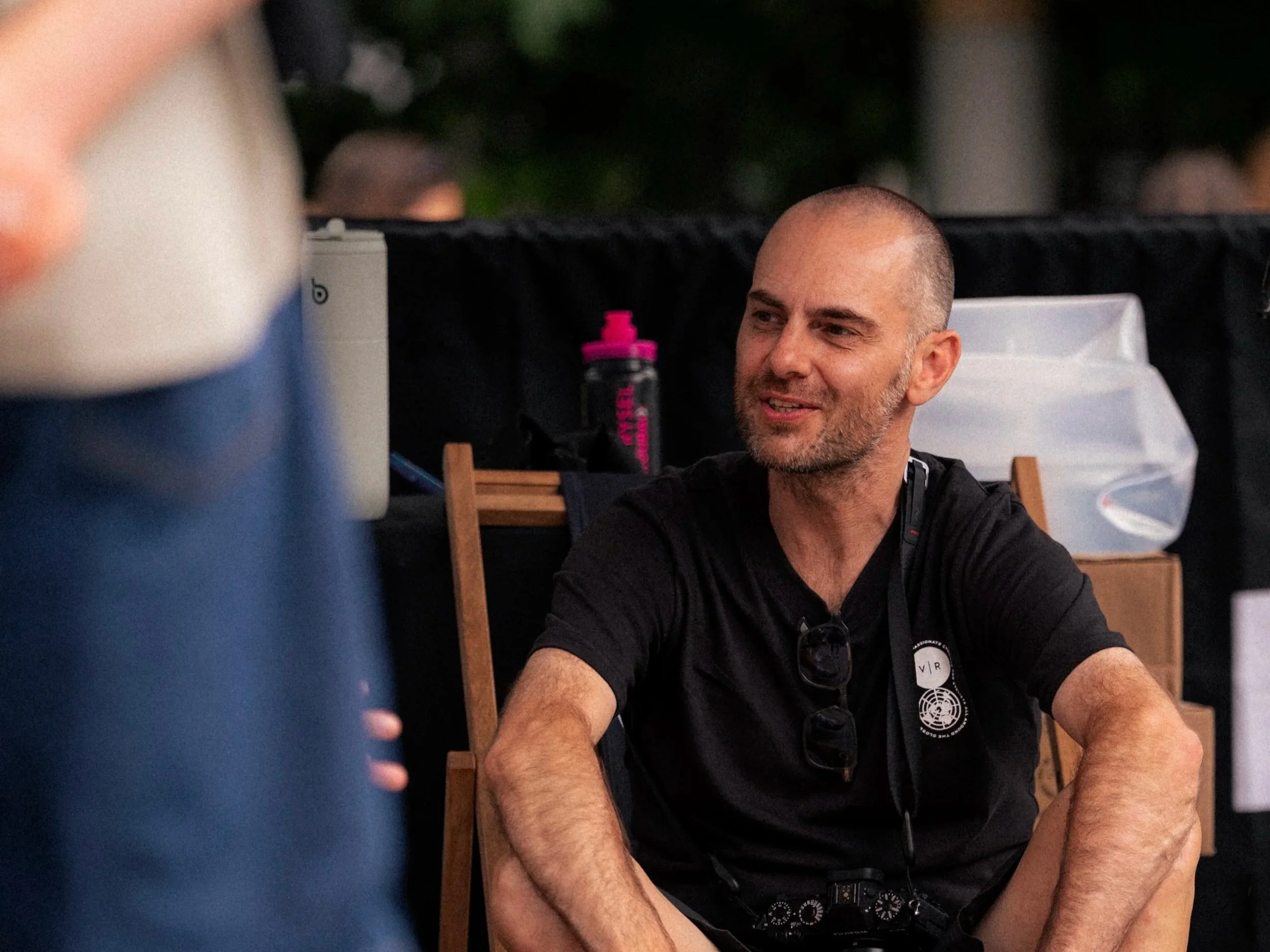 A man with a shaved head and beard, wearing a black shirt with a logo, sitting outdoors, smiling and talking, with sunglasses hanging from his shirt. There are plastic storage containers and a pink water bottle in the background.