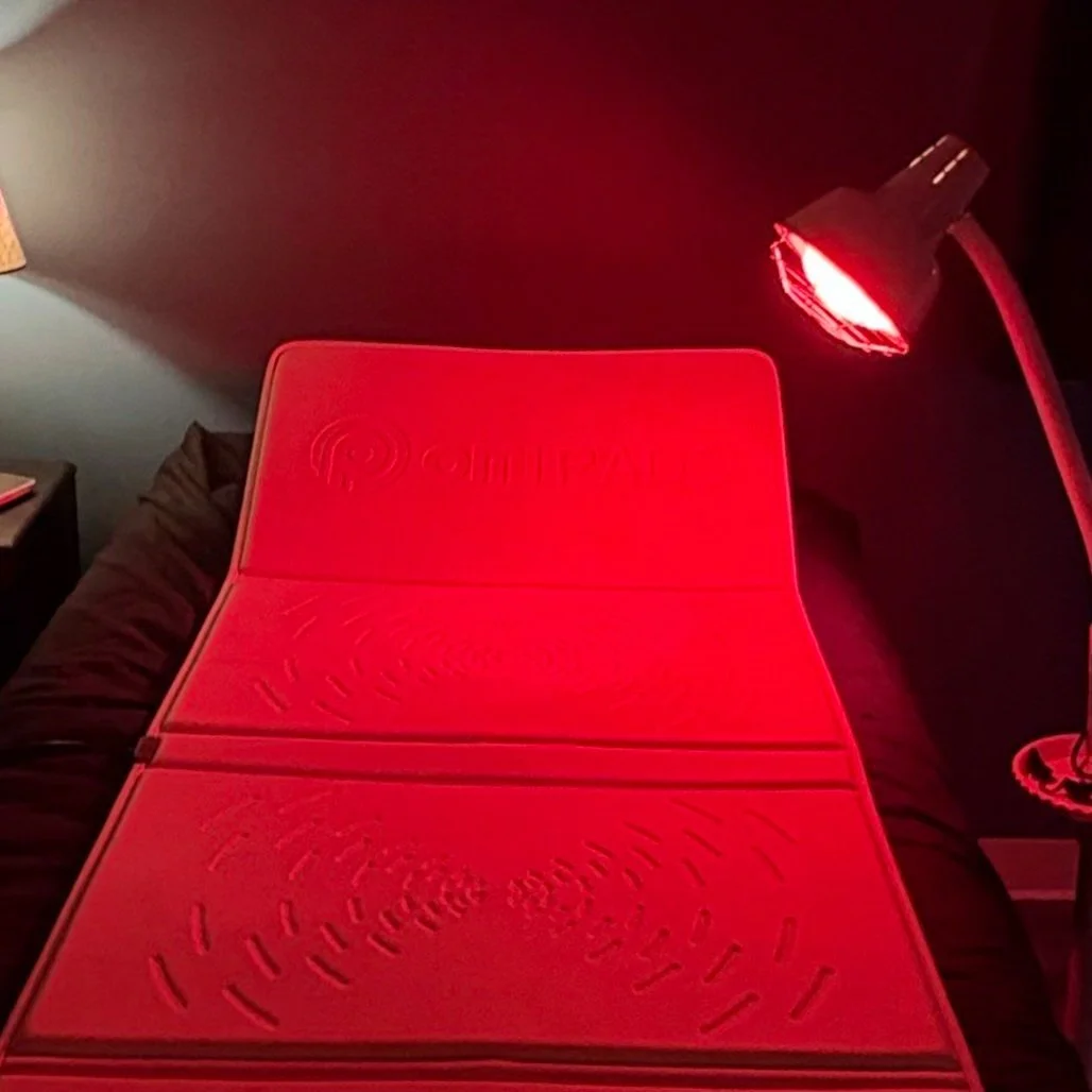 A red-tinted stacked mattress with a wireless logo, placed on a bed with dark bedding. A red lamp illuminates the scene, casting red light on the mattress and surrounding area.