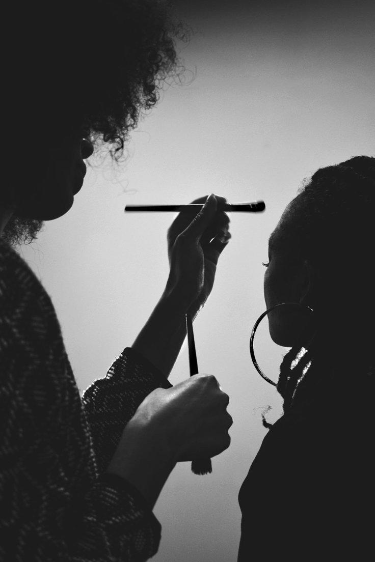 A person applying makeup to another person in silhouette, with a makeup brush visible.