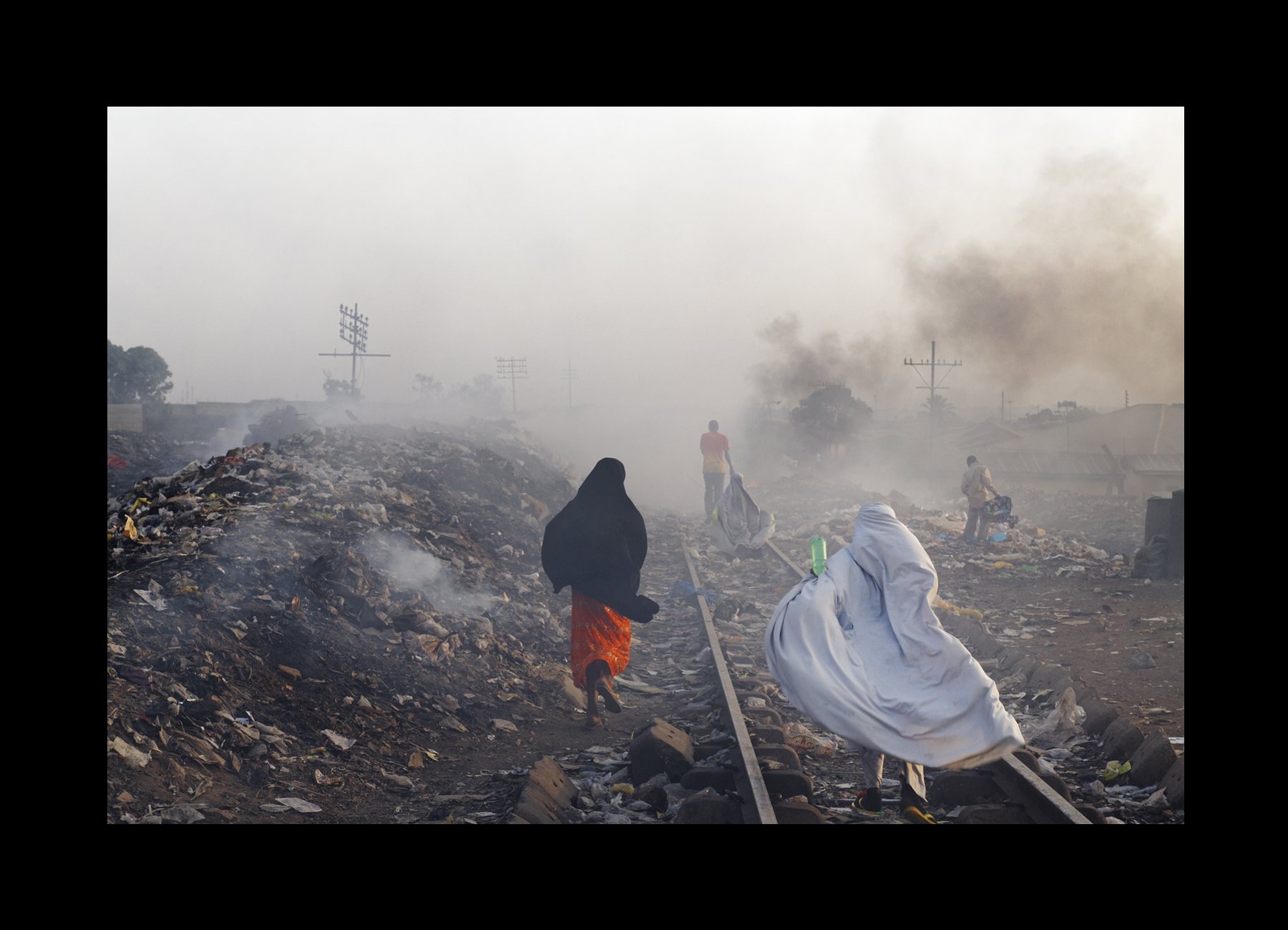    Kaduna, Nigeria, 2013    Locals go about their routines, stepping over garbage heaps that burn along the railroad tracks.    