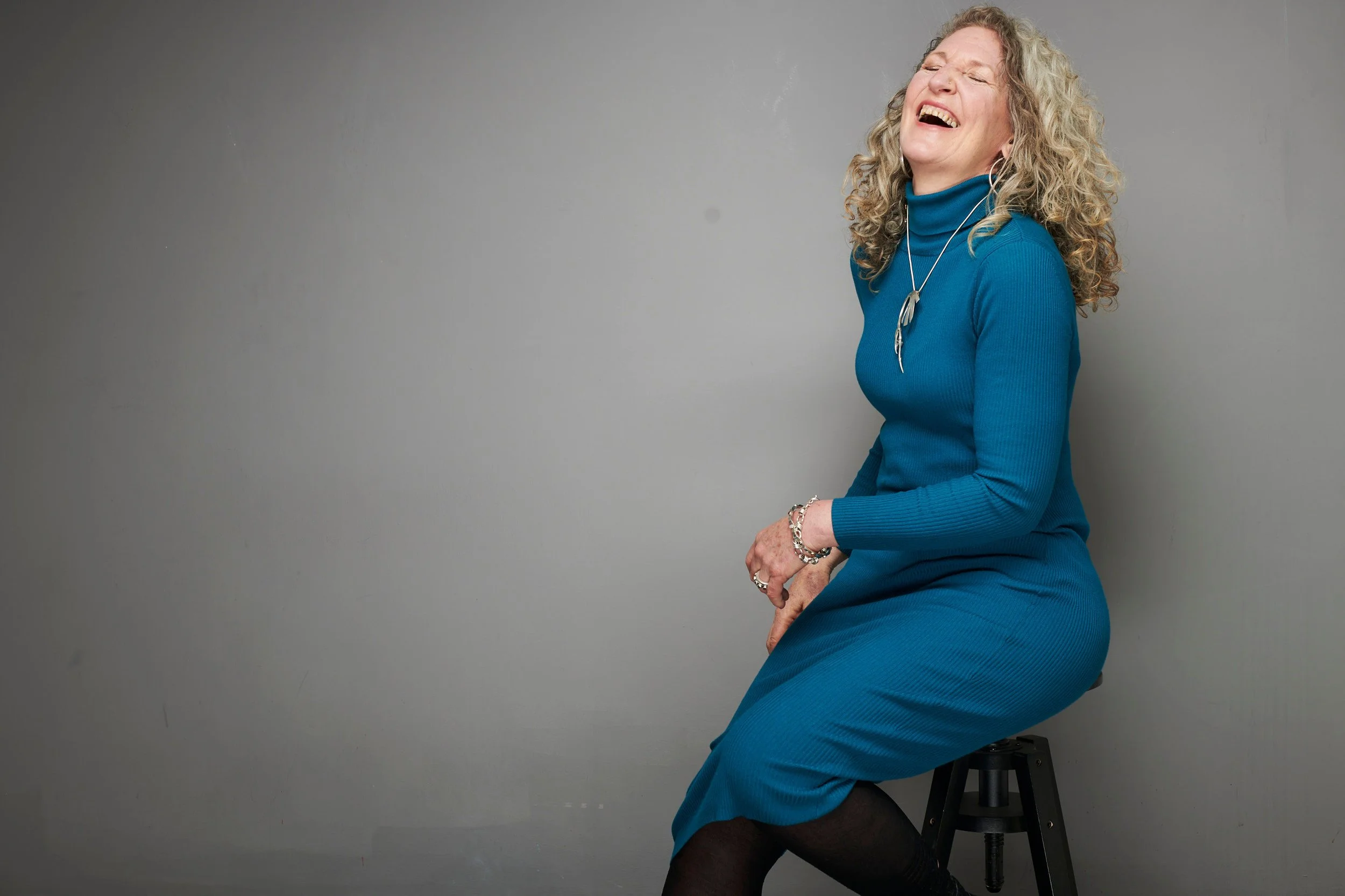Woman in a teal dress laughing as she sits on a stool against a grey backdrop