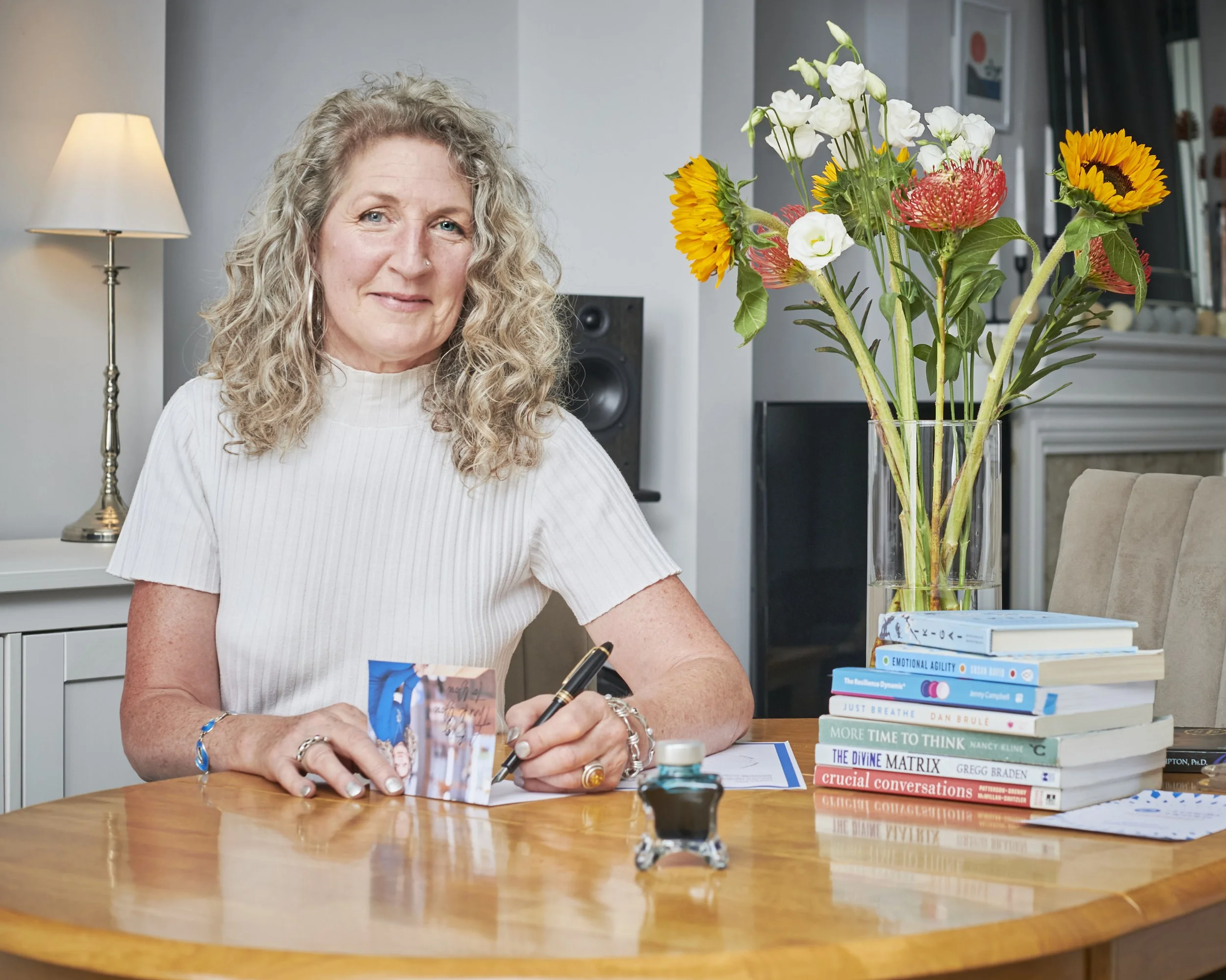 Woman in a white top sitting at a wooden table with books and a vase of sunflowers