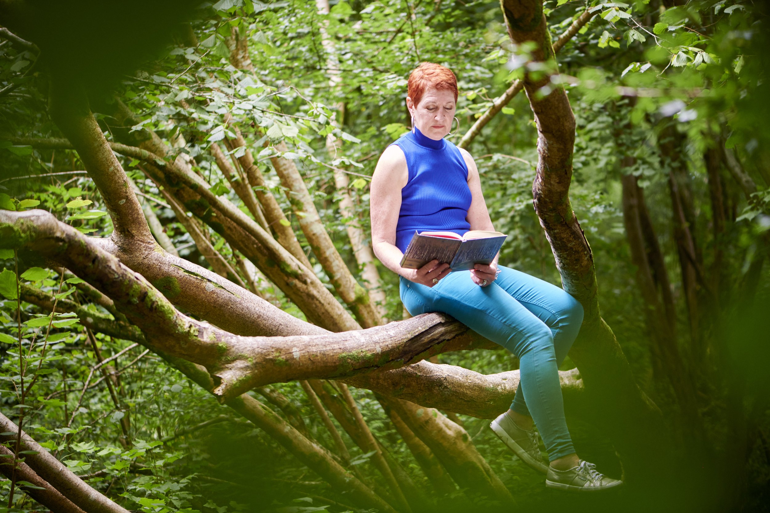 Woman in a blue top reading a book while sitting on a tree branch in woodland