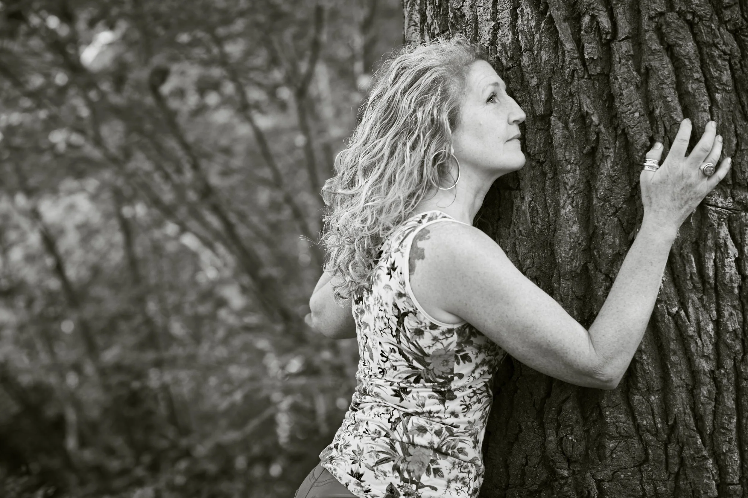 Black and white photo of a woman with her hands on a tree trunk, looking up