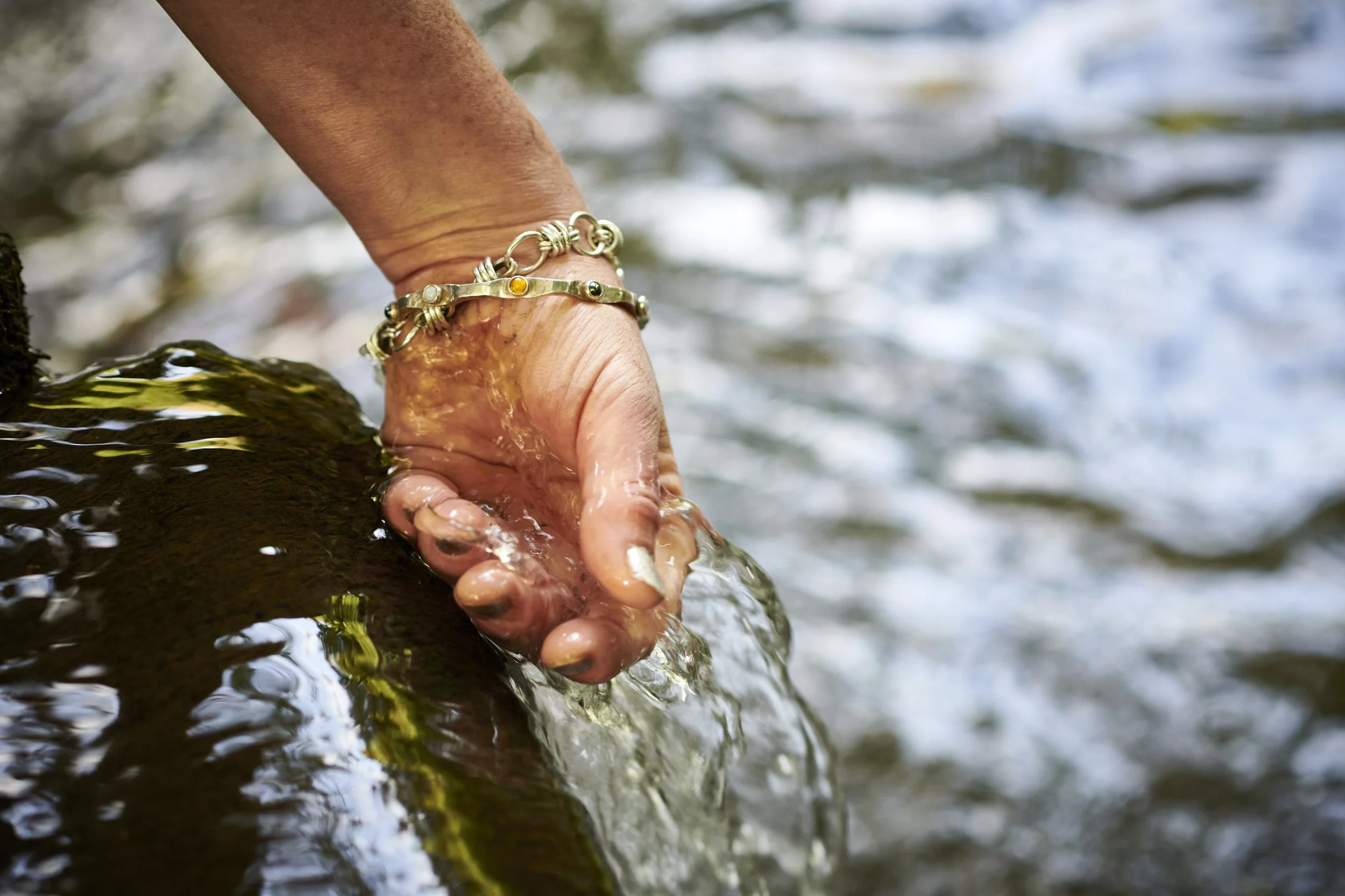 Close-up of a hand wearing a gold bracelet, cupping water from a stream
