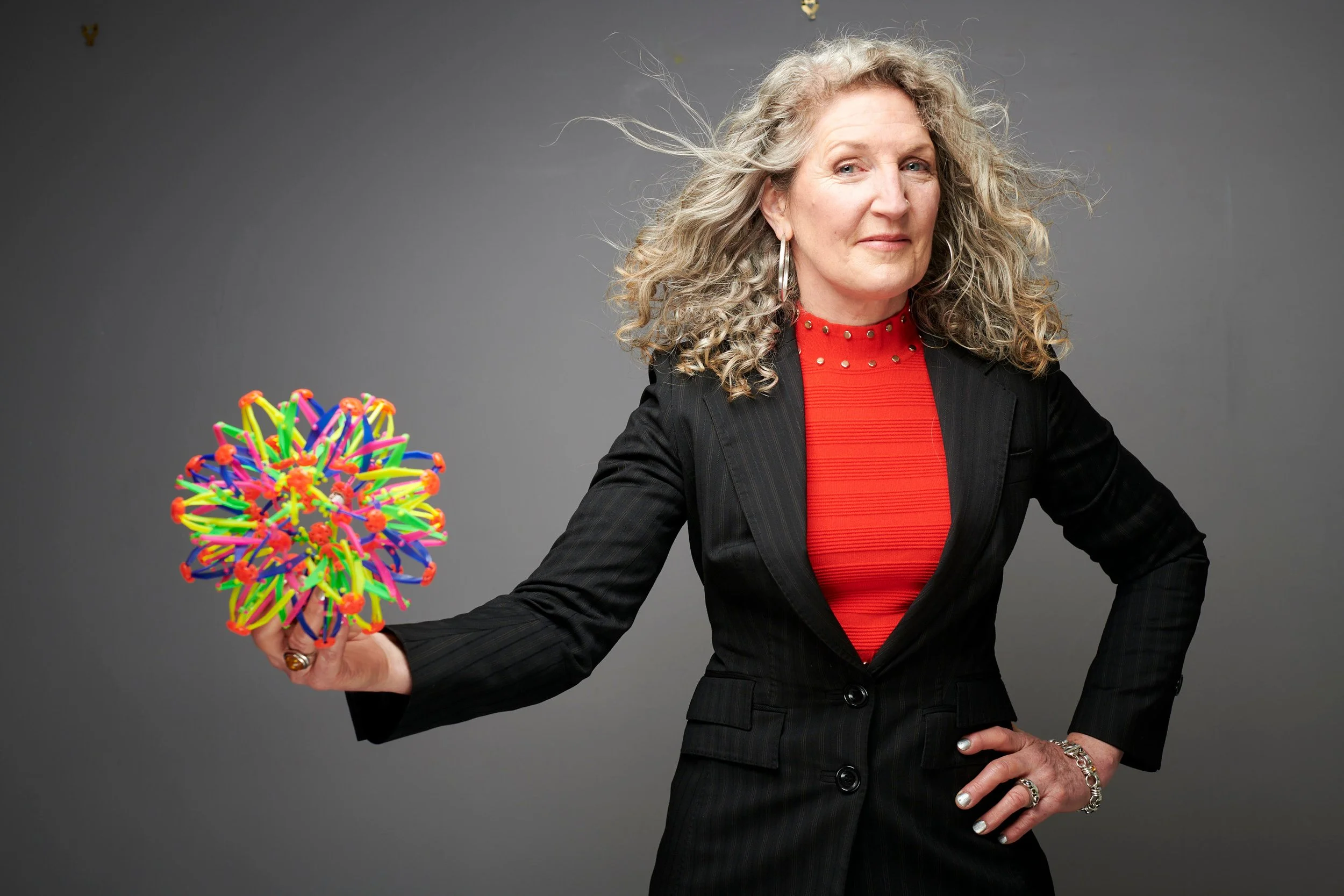 Woman in a black blazer and red top holding out a rainbow-coloured expanding sphere toy