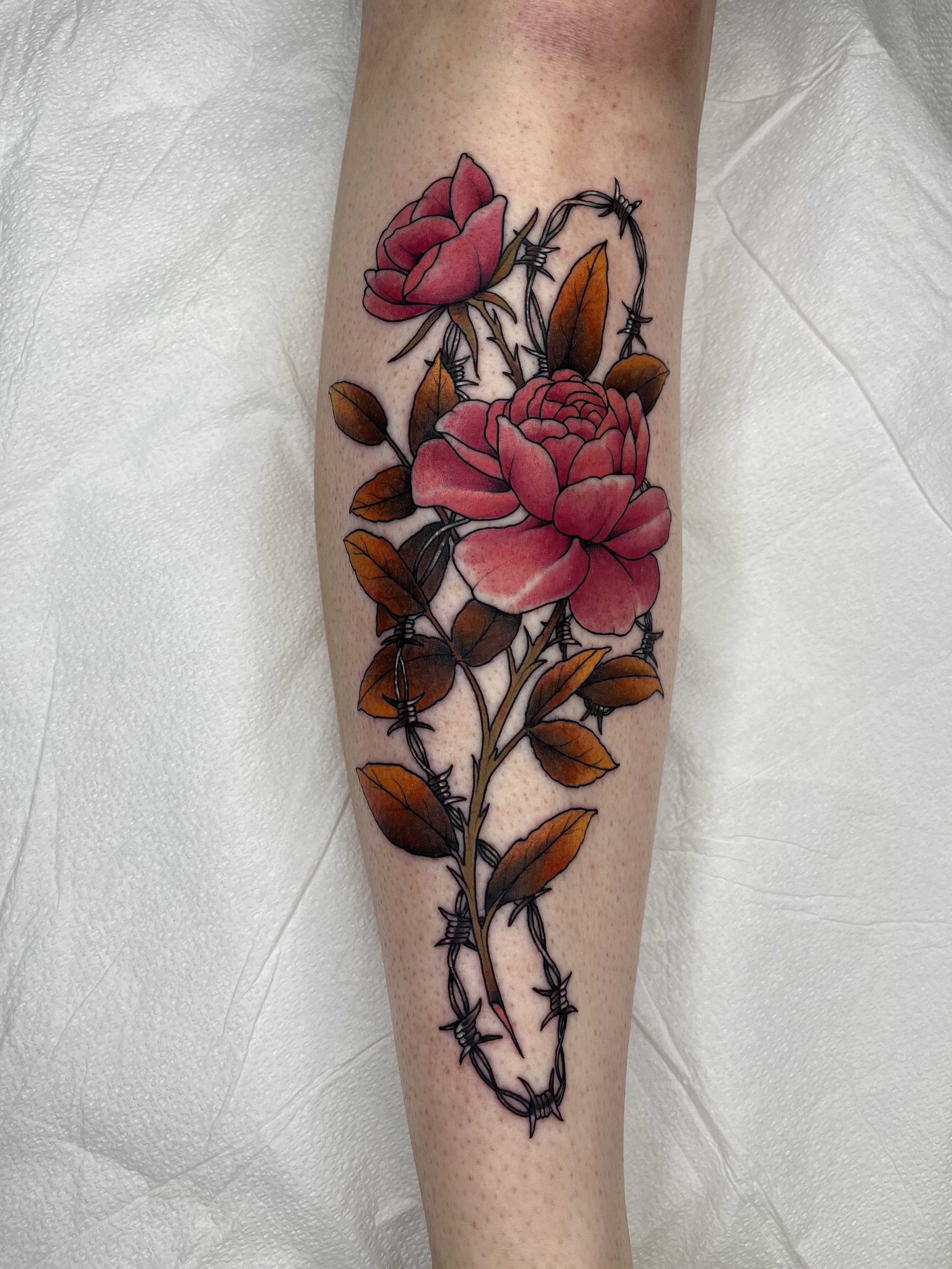 A tattoo of two pink roses, with brown leaves, surrounded by barbed wire, on a person's forearm.