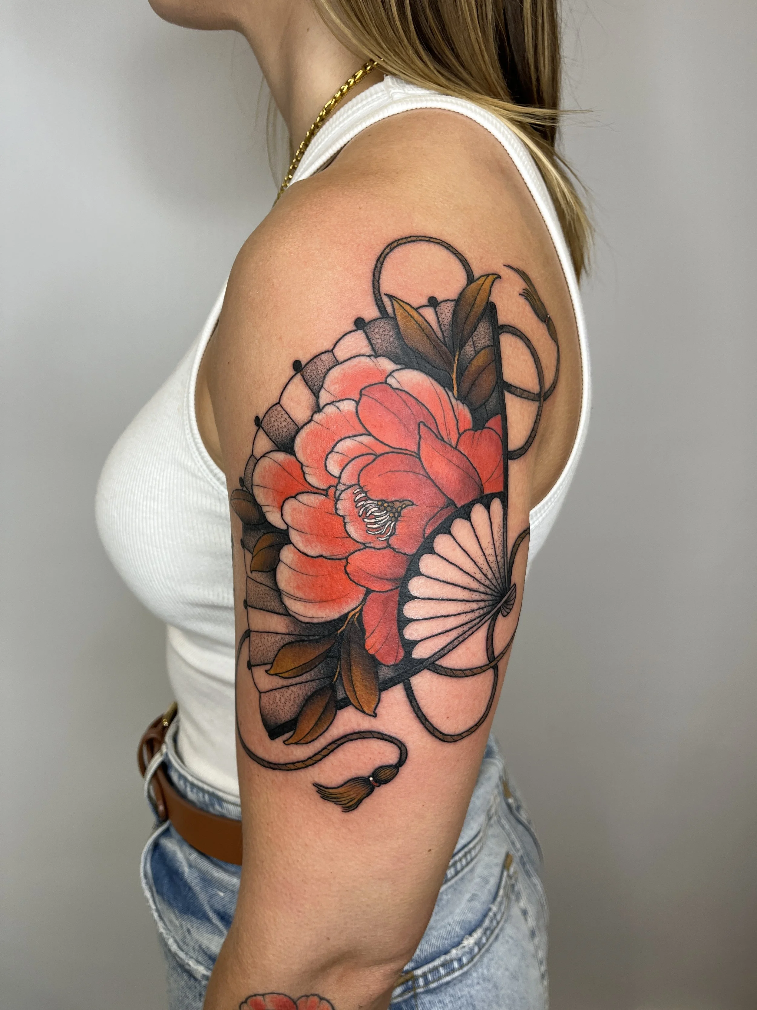 Tattoo of a large red flower with green leaves and a fan on a woman's upper arm, outlined in bold black lines.