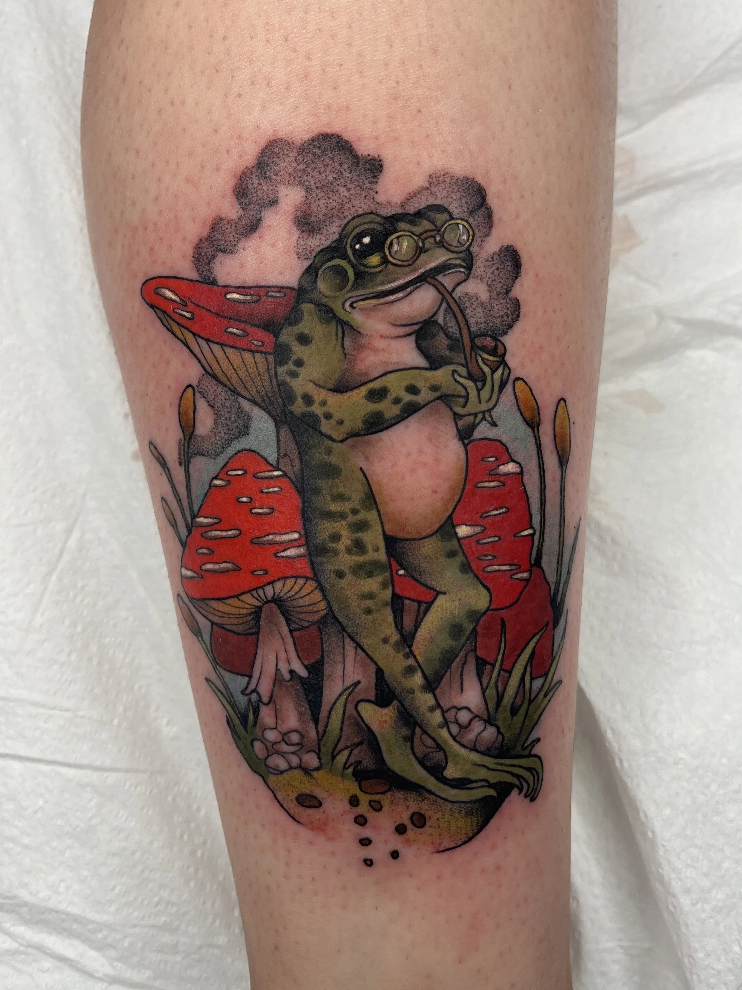 Colorful tattoo of a frog with glasses sitting on red mushrooms, drinking from a pipe, with smoke in the background.