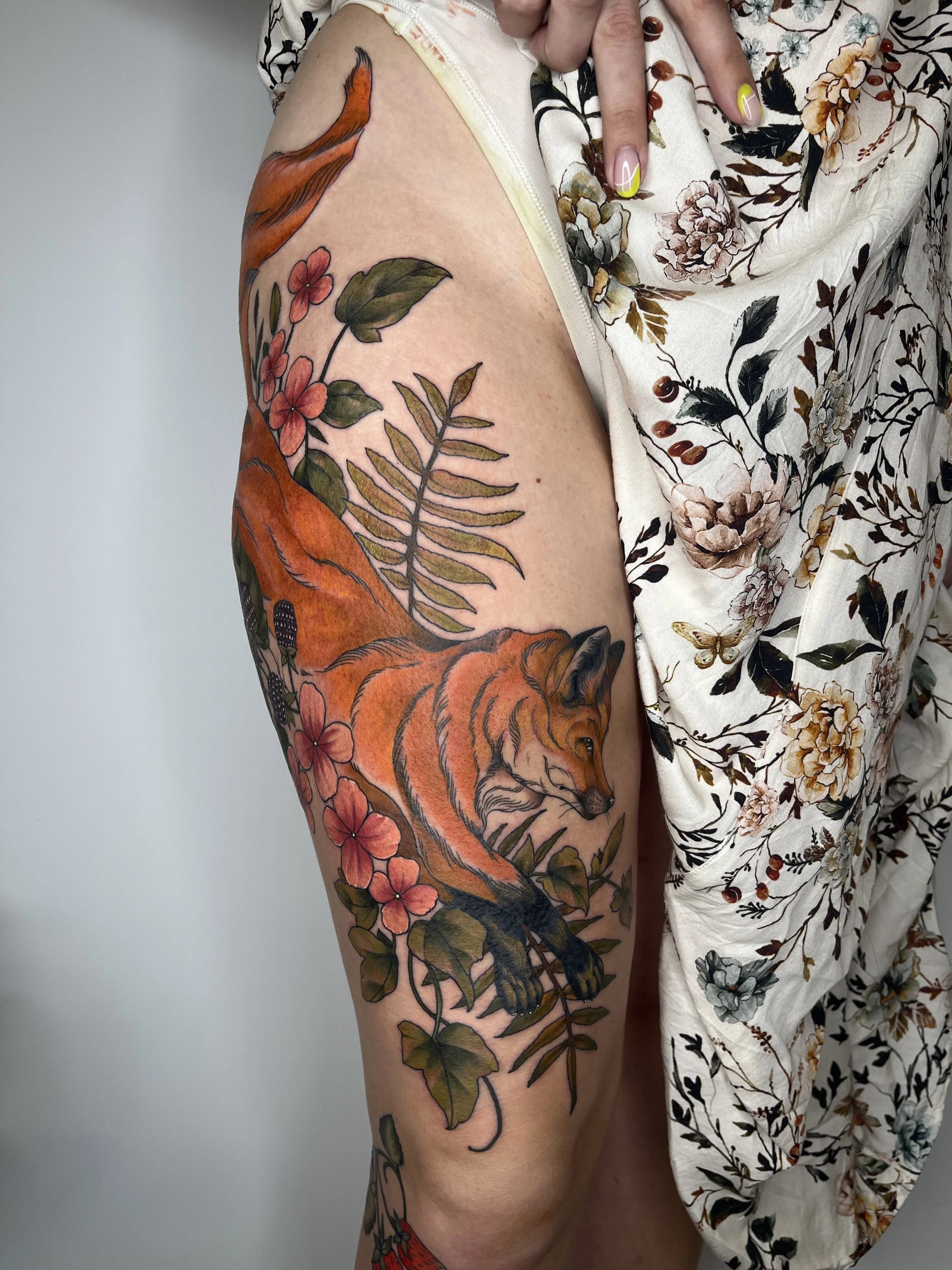 Tattoo of a fox surrounded by flowers and leaves on a person's thigh.