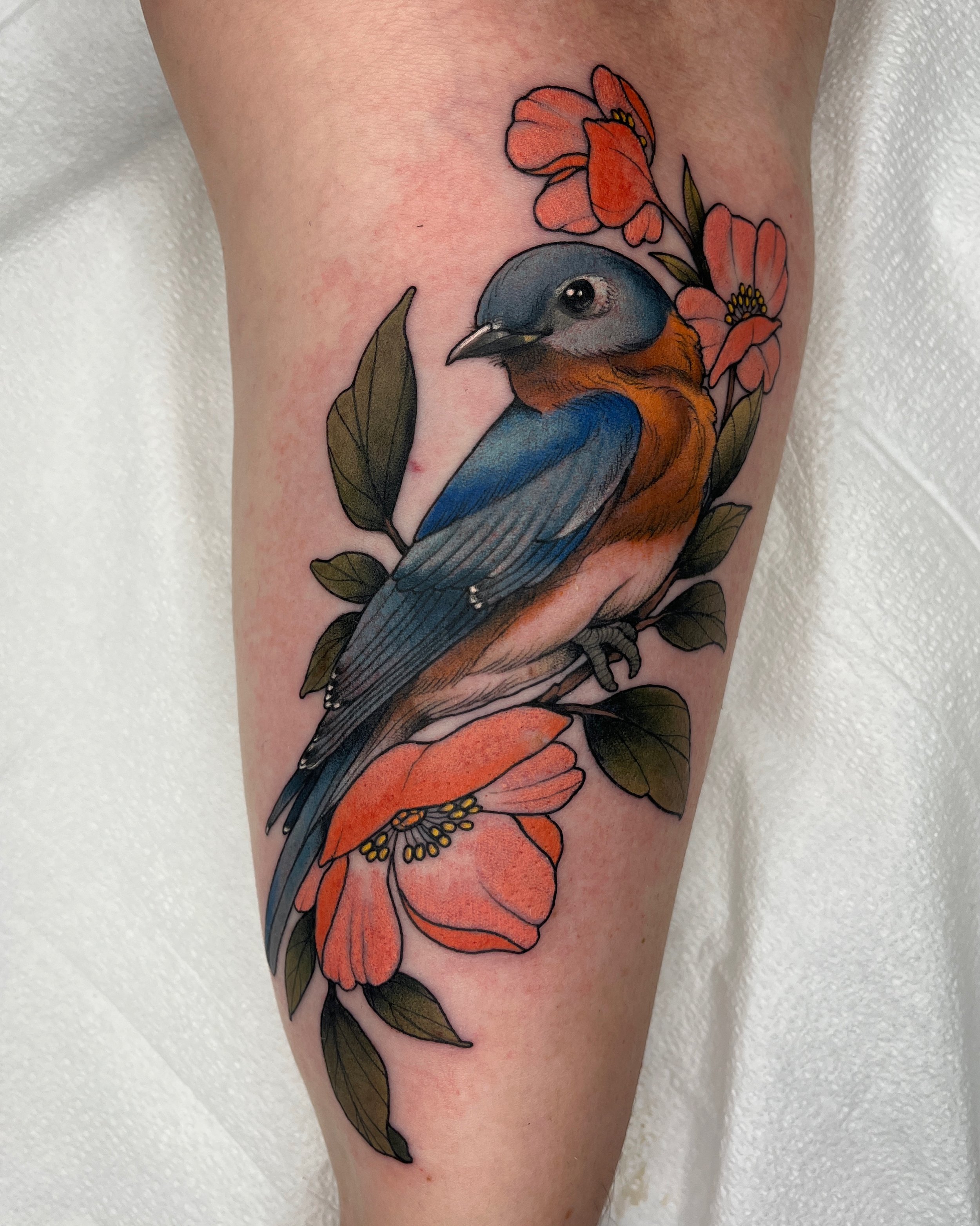 Colored tattoo of a bird with blue, orange, and gray feathers perched among orange flowers and green leaves.