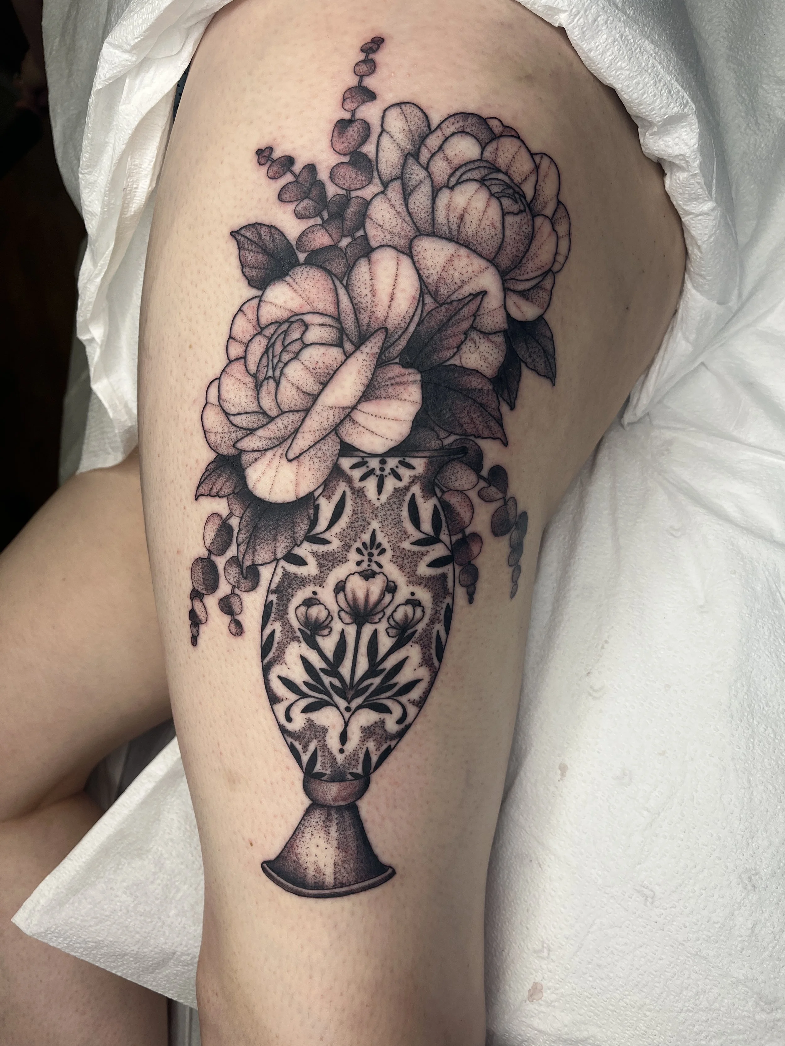 A black and gray tattoo of a bouquet of flowers in a decorative vase on a person's upper arm.