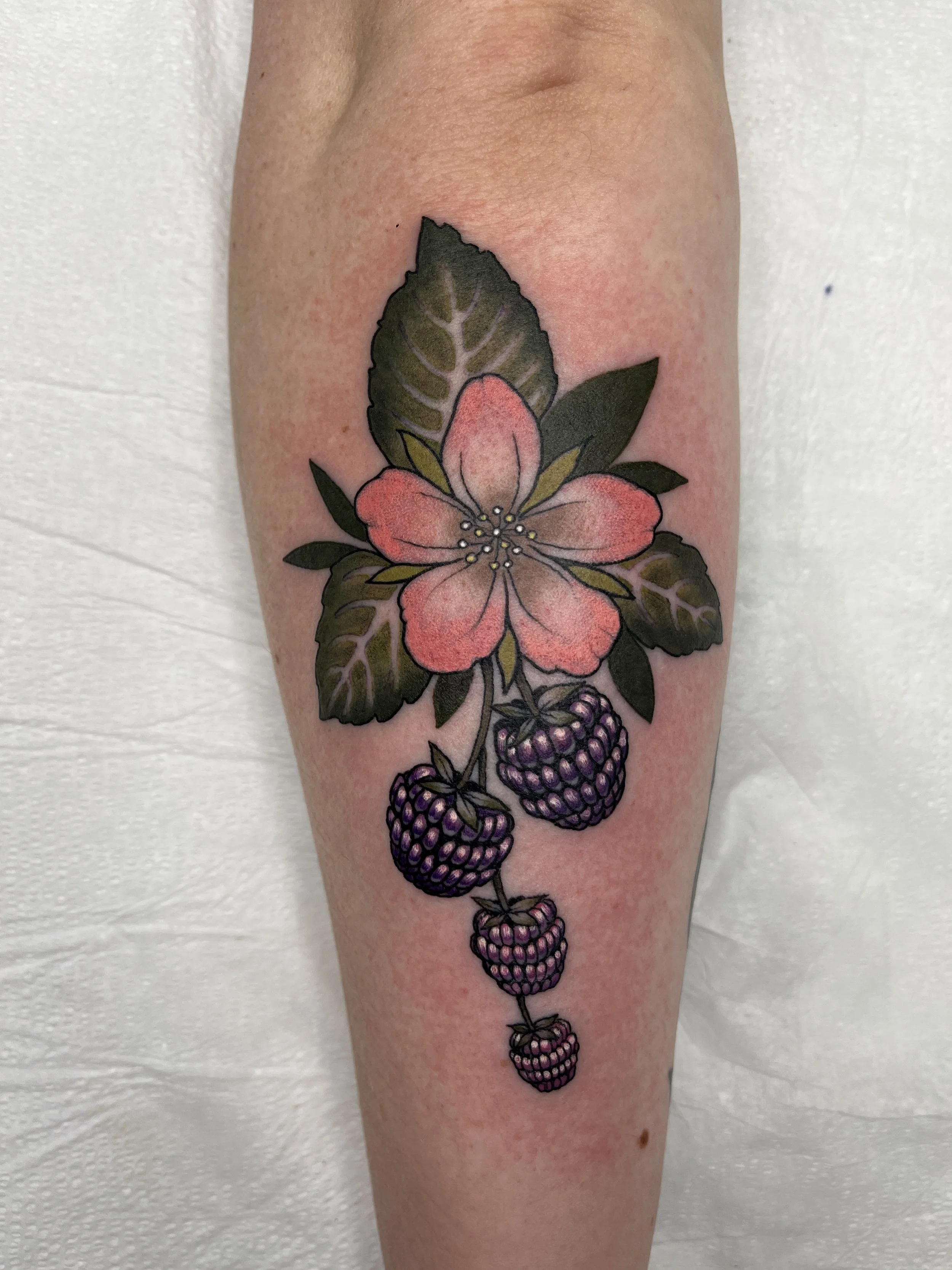 Tattoo of a pink flower with green leaves and purple blackberries on a person's leg.