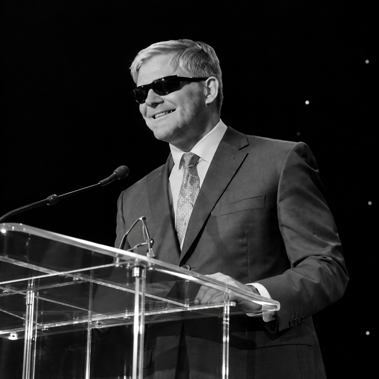Man wearing sunglasses speaking behind clear podium.png