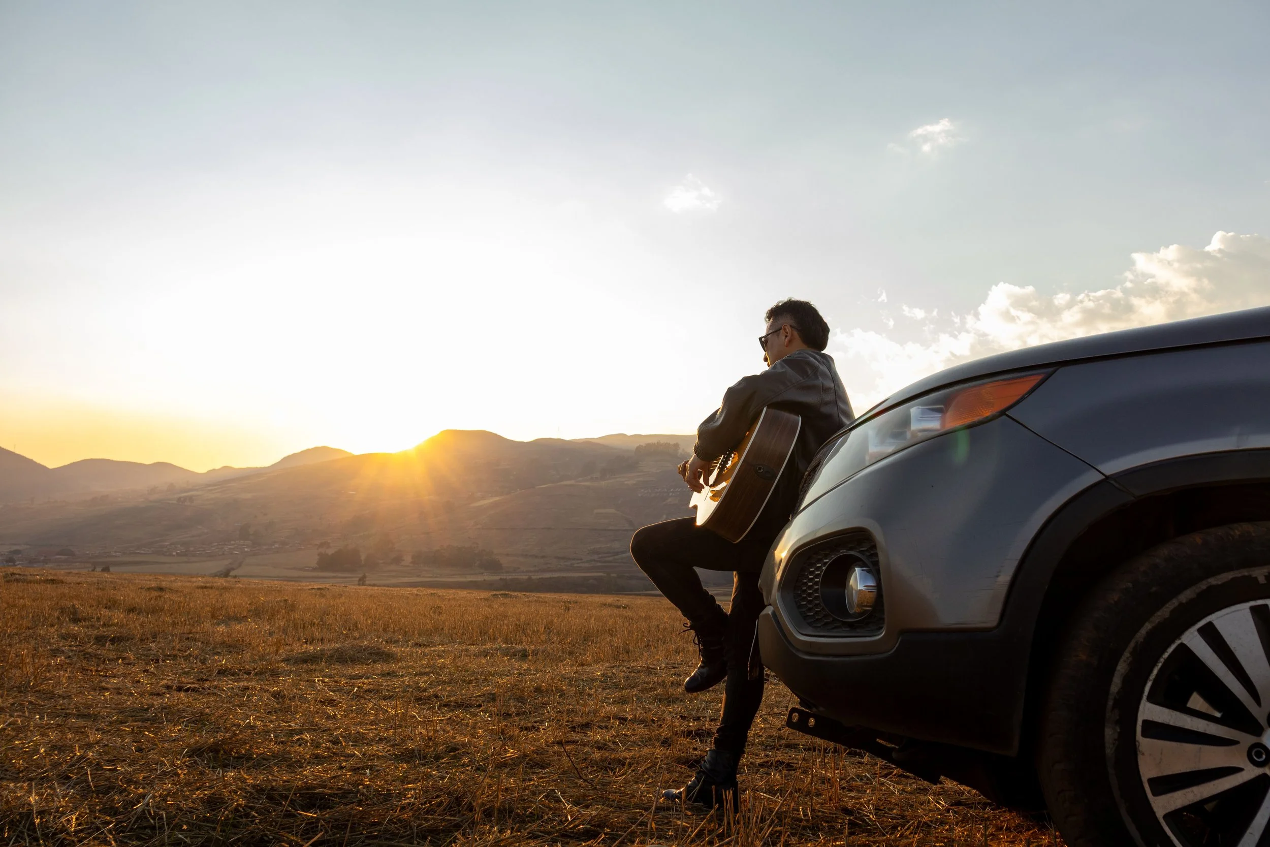 Driver relaxing with a guitar beside his car at sunset after getting high-risk car insurance in California