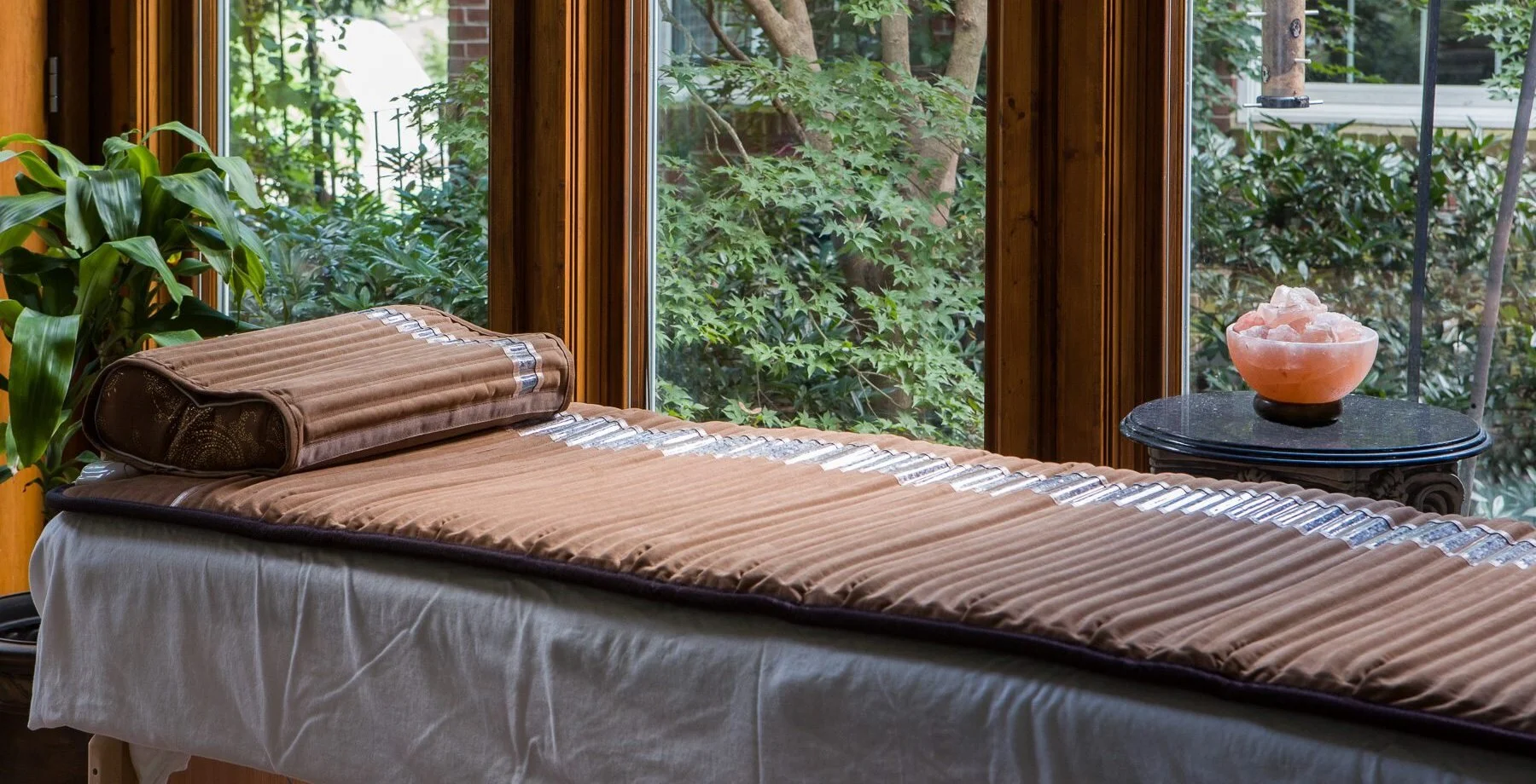 A massage table covered with a beige blanket and a matching pillow, situated near large windows with a view of green foliage outside. There is a small black table with a pink Himalayan salt lamp on it to the right side, and a green plant is visible o