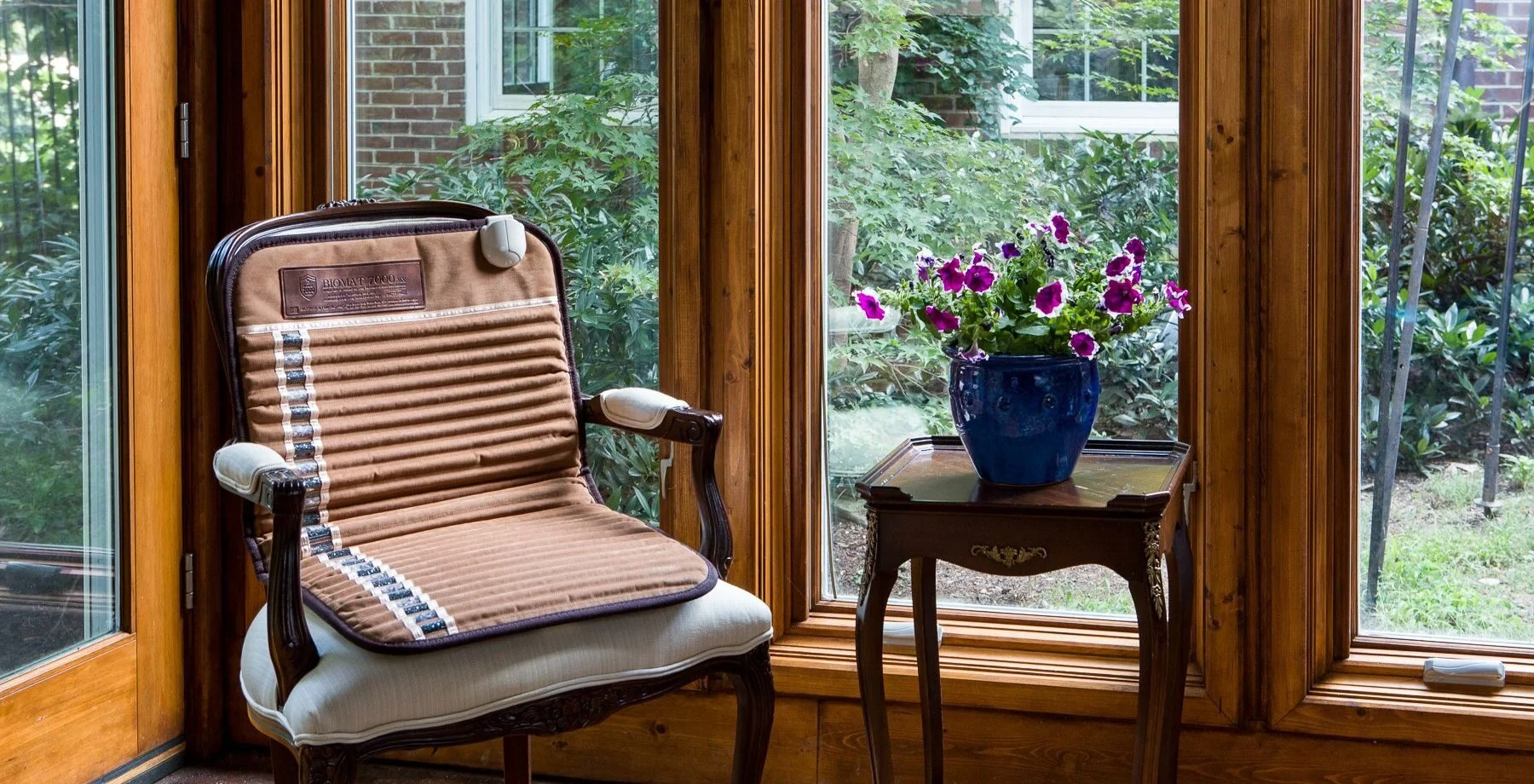 A cozy sunroom with a wooden framed glass door, a vintage wooden chair with a pillow, a small wooden table with decorative details, and a blue ceramic pot with purple and white petunias outside.
