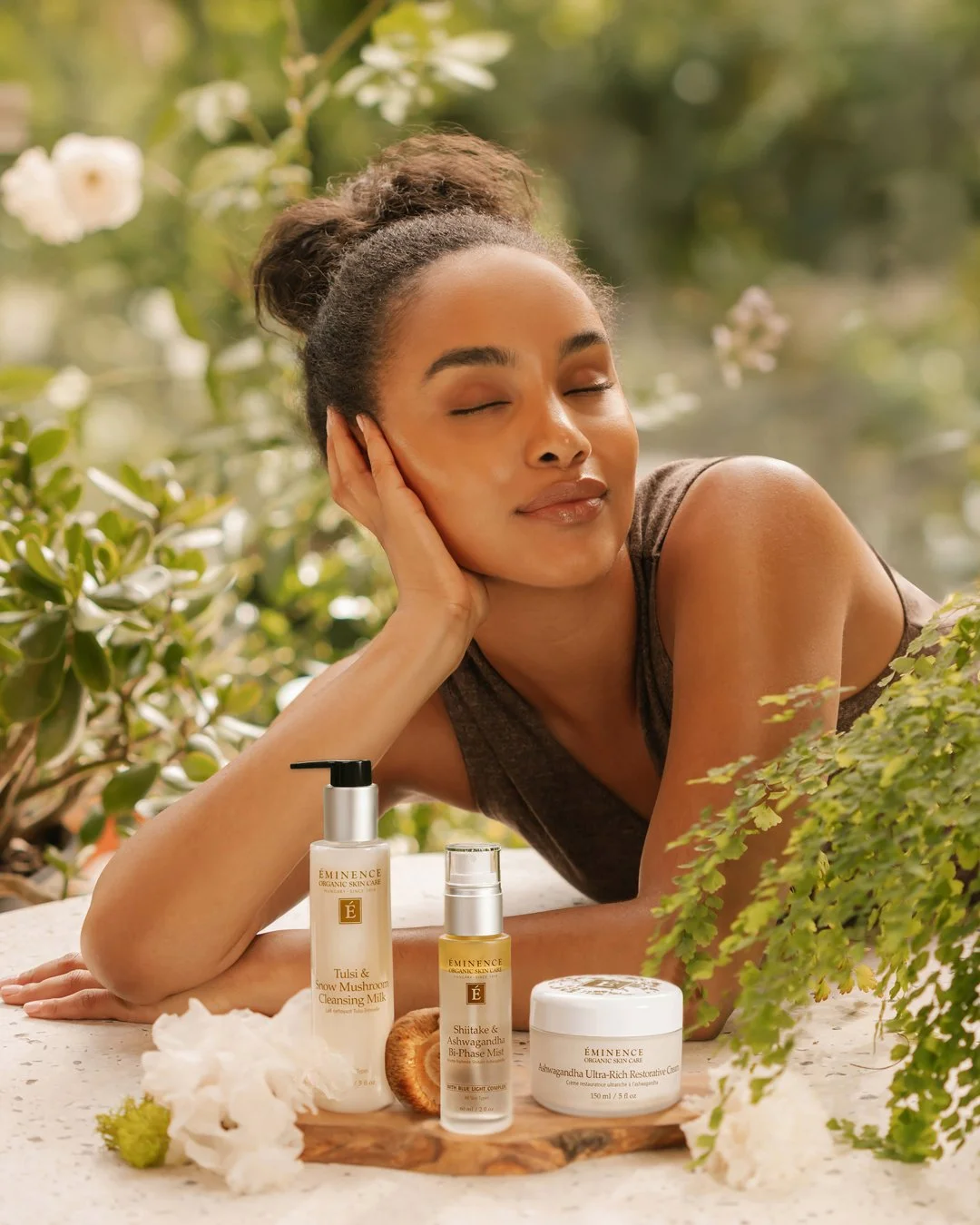 A woman with closed eyes resting her face on her hand outdoors surrounded by greenery and flowers, with skincare products in front of her on a wooden surface.