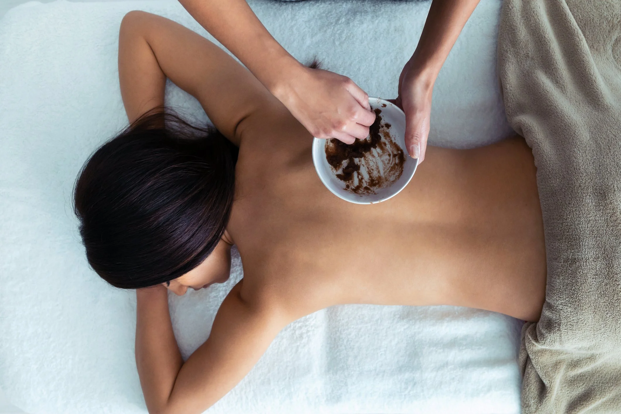 A woman lying on her stomach receiving a massage, with a bowl of herbal massage cream on her back.